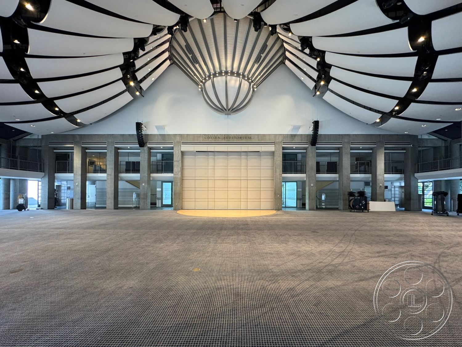Museum 2 - interior space, exposed concrete walls, textured carpet flooring, glass entrance doors, metal railing accents, vaulted ceiling with curved panels, central lighting fixture, open auditorium layout, columned structural supports, multiple large windows