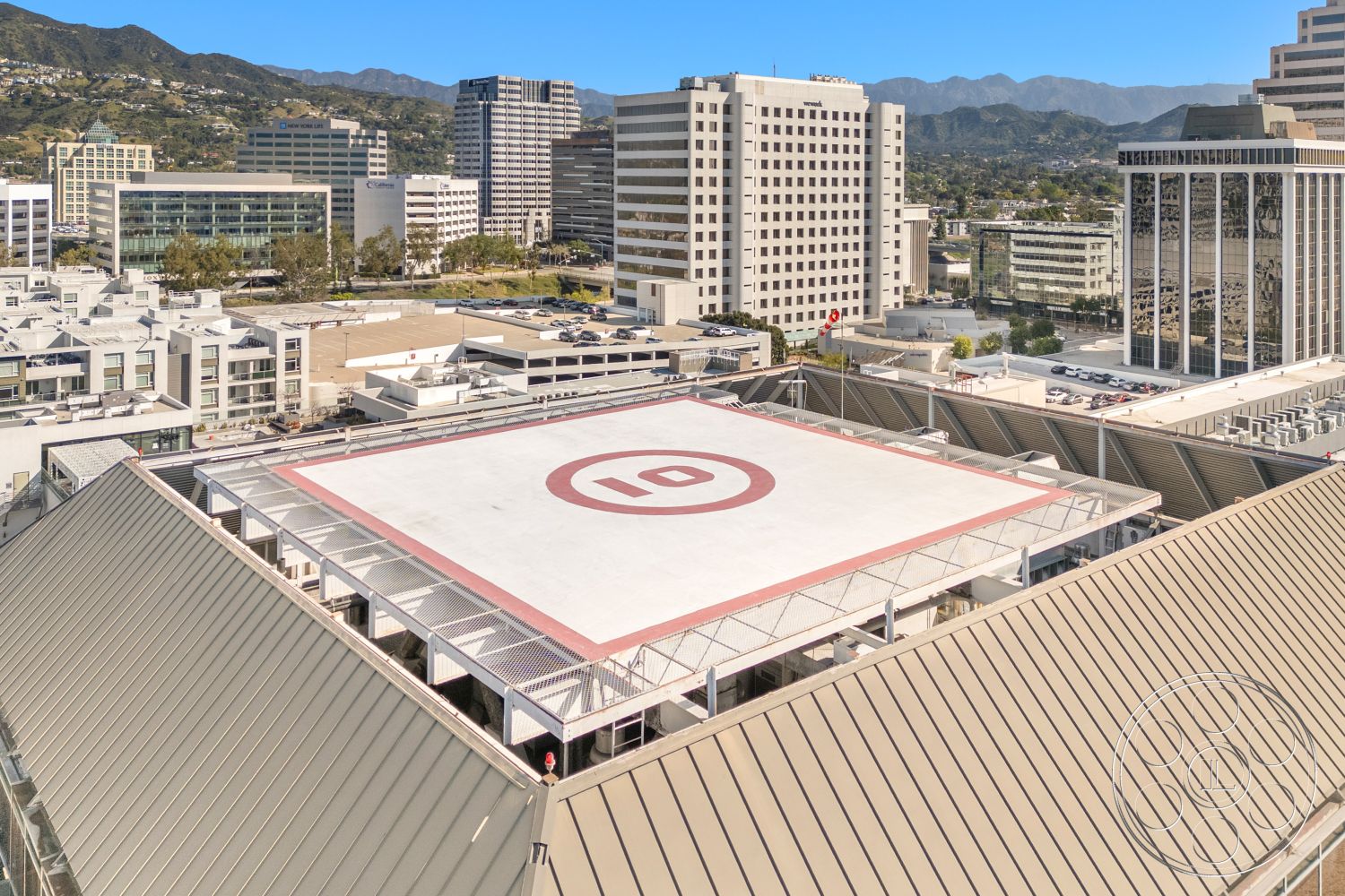Rooftop-52 - exterior, urban environment, flat rooftop, concrete parking structure, mountain view in background, metal roof with angled profile, glass building facades, commercial district skyline, helipad with red outline