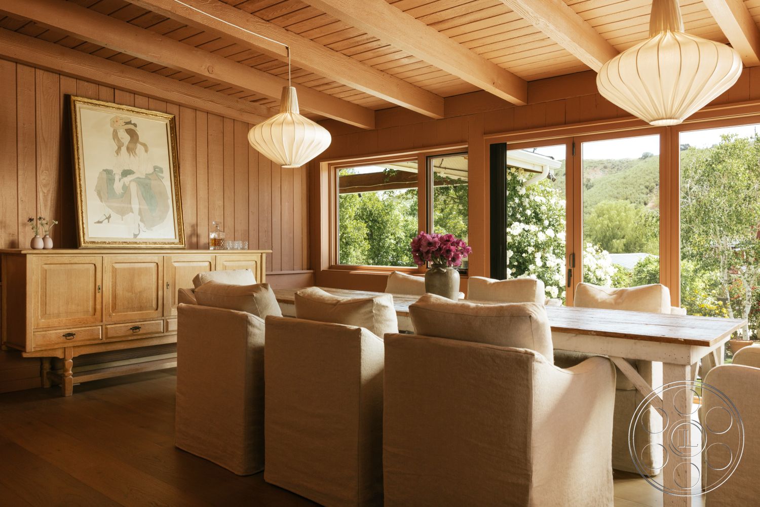 Modern-Farmhouse 20 - upholstered dining chairs, wooden sideboard, neutral color scheme, light wood dining table, pendant lighting fixture, wood beam ceiling, exposed wood ceiling, natural light flooding the room, large windows with nature view, textured wood panel walls, artwork with gold frame