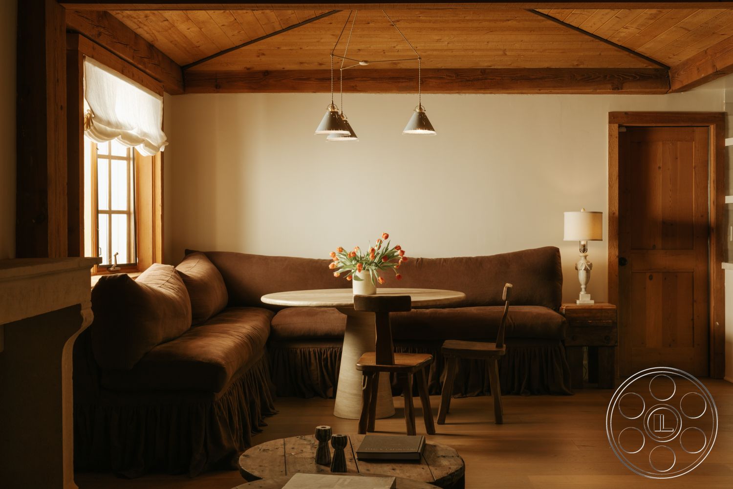 Modern-Farmhouse 20 - hardwood flooring, modern pendant lighting, natural light from window, wooden dining chairs, exposed wood ceiling beams, wood paneling walls, brown upholstered sectional sofa, round dining table with pedestal base, glass table centerpiece with flowers, rustic style decor, wooden door with paneling