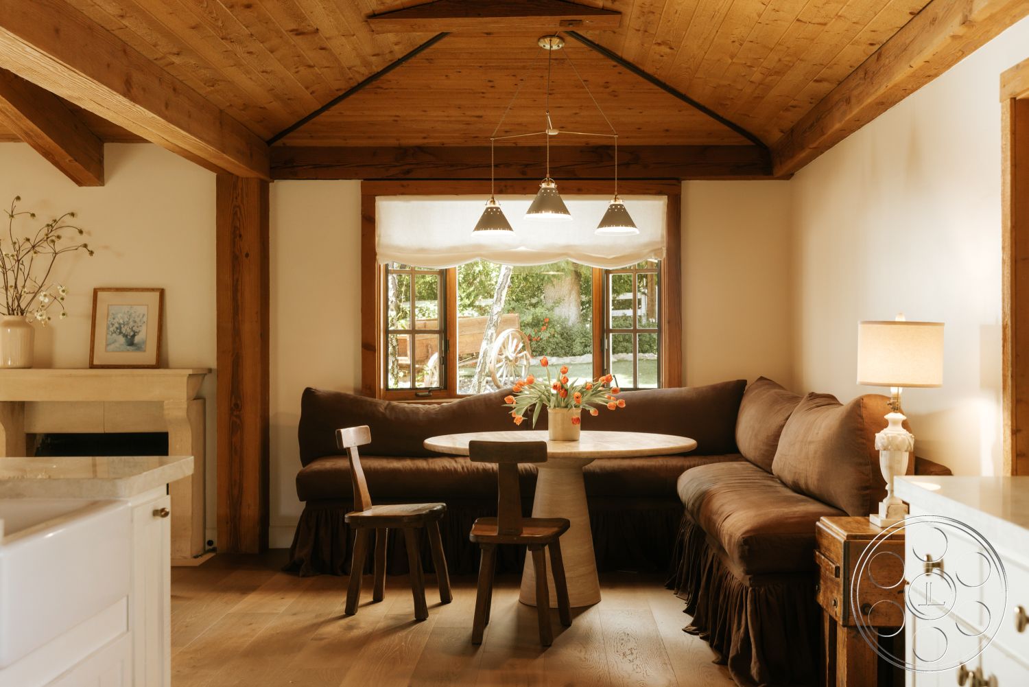 Modern-Farmhouse 20 - hardwood flooring, round dining table, natural wood paneling, wooden dining chairs, neutral painted walls, decorative vase with flowers, exposed wood beam ceiling, stone fireplace surround, minimalist light fixtures, fabric-upholstered sofa, large window with views, artisan wooden side table