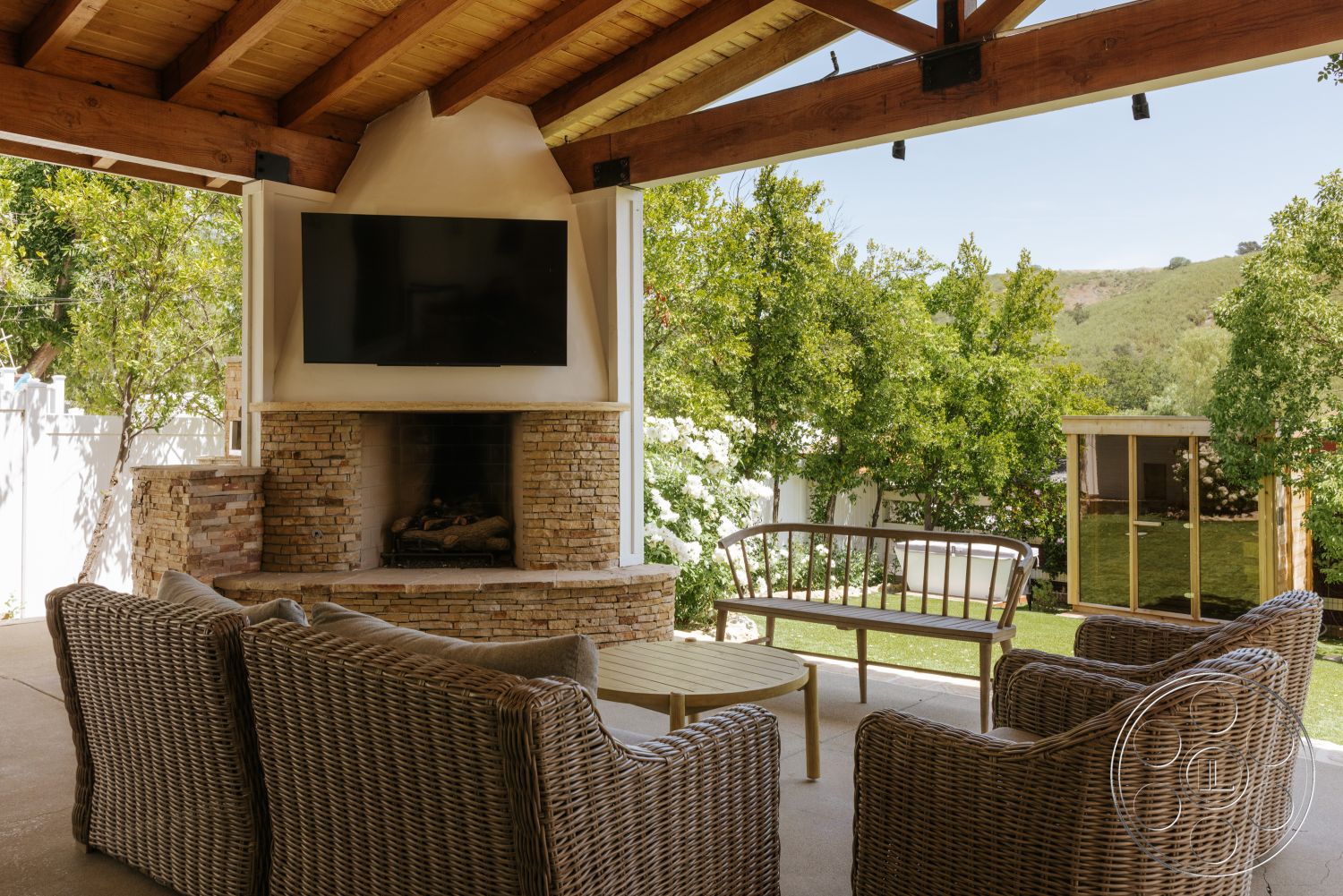 Modern-Farmhouse 20 - exterior, landscaped garden view, outdoor entertainment area, natural light-filled space, wood beam ceiling, concrete patio floor, stacked stone fireplace surround, woven wicker seating, flat-screen TV mounted, open-air shelter structure