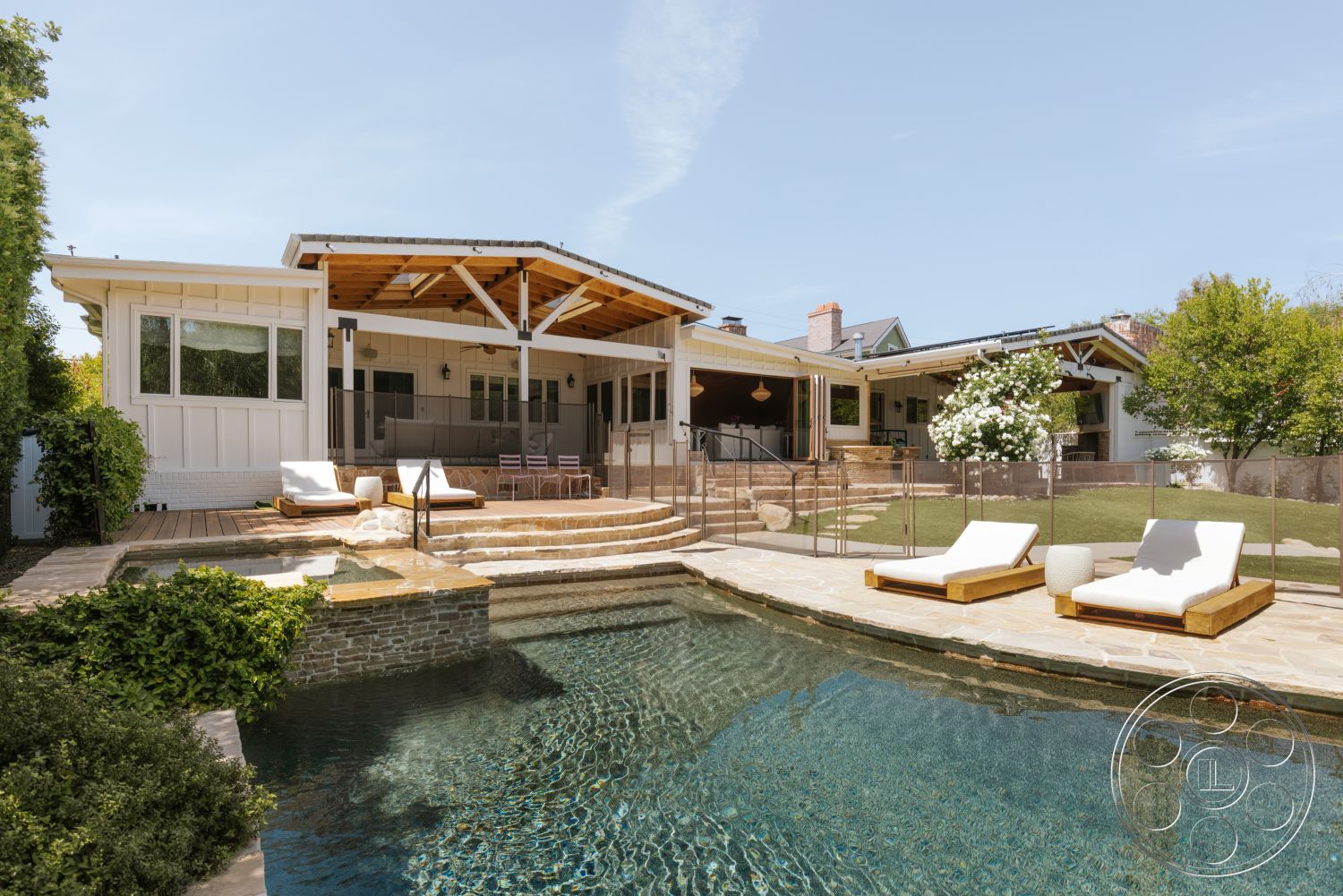 Modern-Farmhouse 20 - exterior, sliding glass doors, surrounding greenery, infinity pool, wooden lounge chairs, sunny backyard, stone pool deck, modern ranch-style home, brick fireplace surround, wood beam ceiling, white painted walls, stone patio flooring, landscaped garden with hedges, vaulted outdoor ceiling, glass pool surround, spacious outdoor patio, sitting area with outdoor furniture