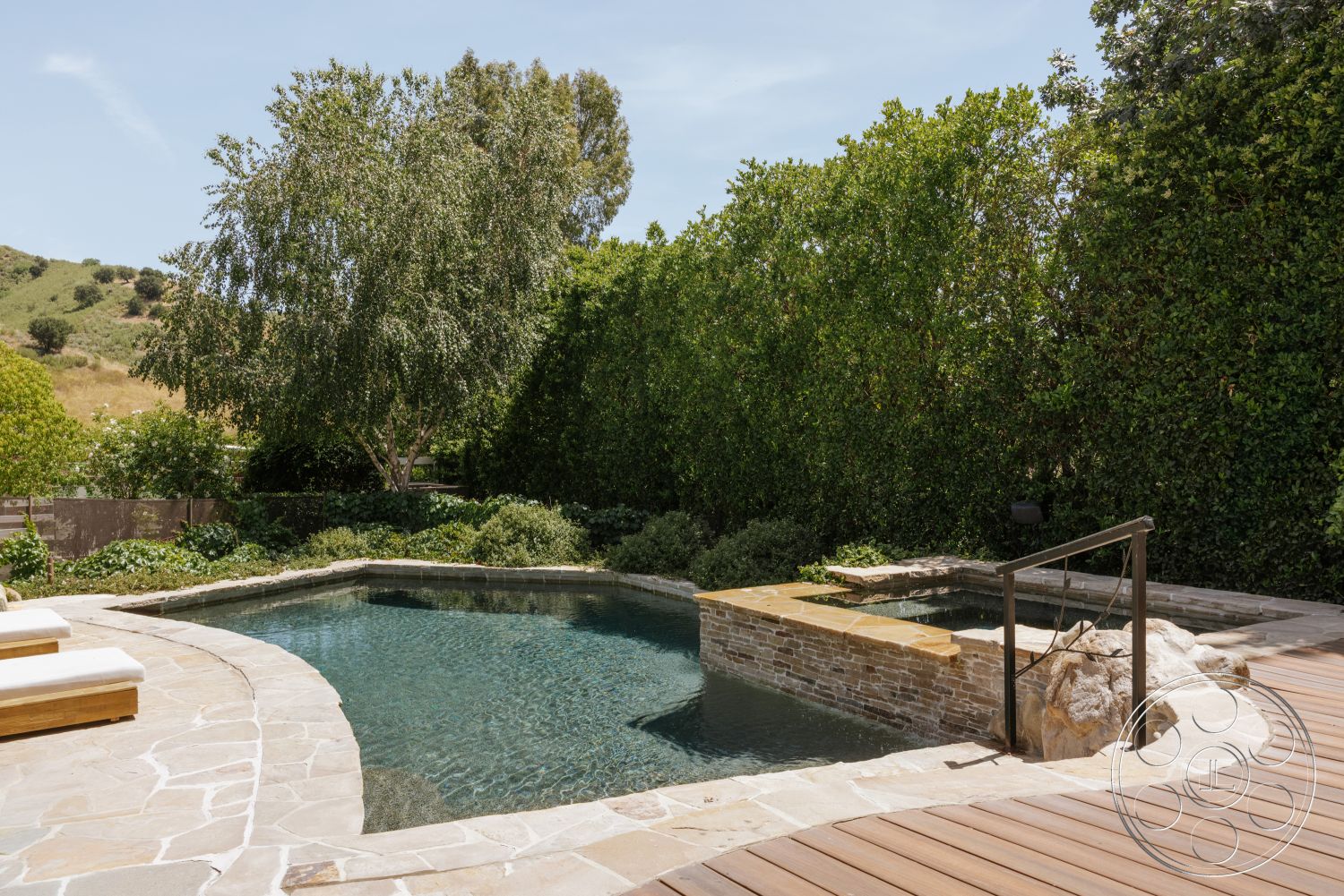 Modern-Farmhouse 20 - exterior, lush greenery, surrounding hillside, paved stone patio, rectangular pool, wooden lounge chairs, stone pool deck, sunny outdoor setting, landscaped garden area, built-in spa, smooth concrete coping, natural stone spa surround