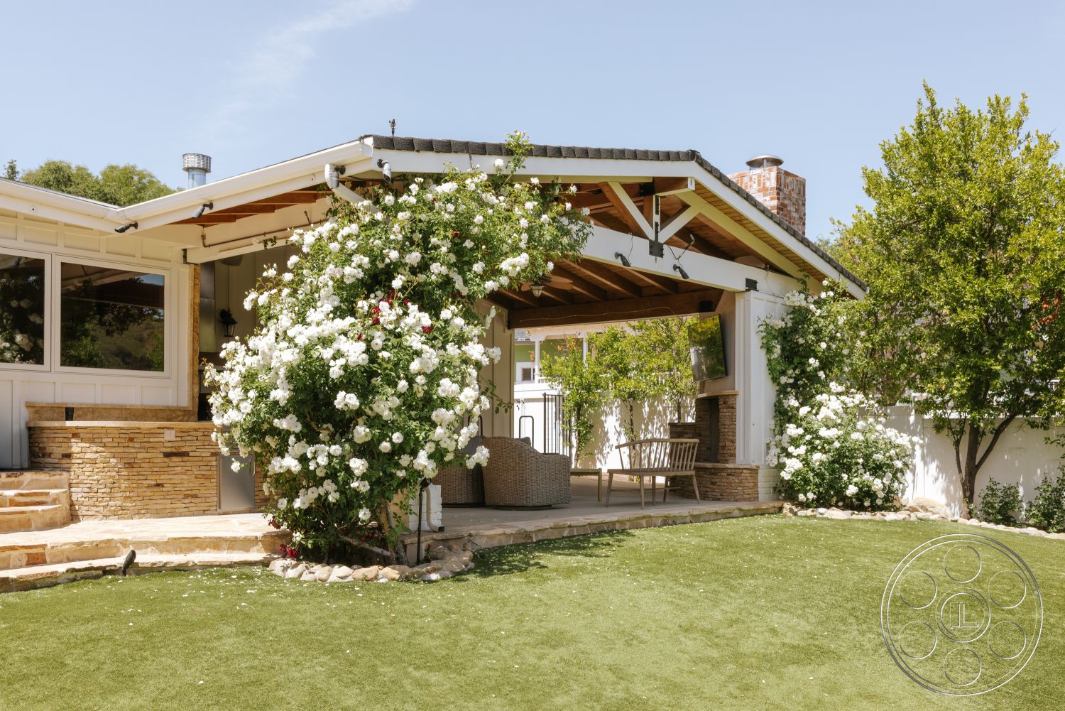 Modern-Farmhouse 20 - large windows, exterior, outdoor seating area, brick chimney, suburban backyard setting, wood beam ceiling, flowering shrubbery, white painted walls, stone fireplace surround, stone patio flooring, lush greenery landscaping, artificial grass lawn, covered patio area