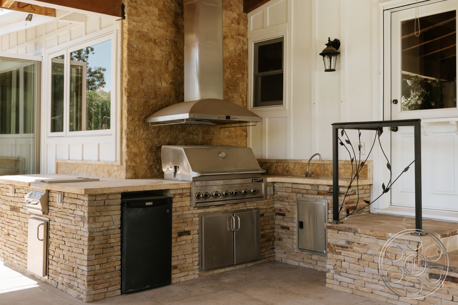 Modern-Farmhouse 20 - outdoor dining space, textured stone backsplash, large window view, vaulted ceiling with exposed wood beams, natural stone feature wall, stainless steel outdoor grill, stone outdoor kitchen countertop, white vertical siding, exterior kitchen area, black outdoor refrigerator, installed outdoor sink, cut stone steps, metal railing with leaf design
