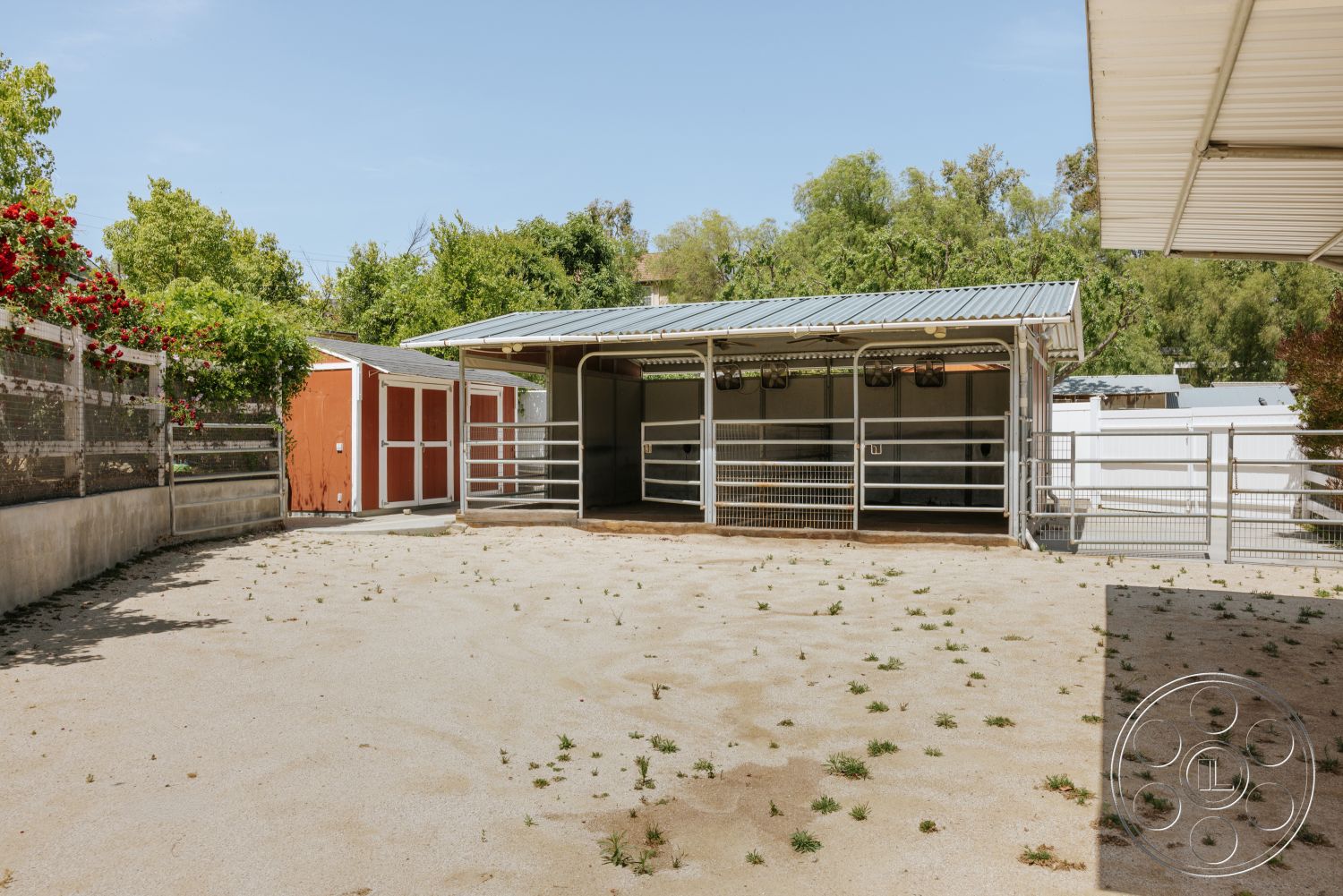Modern-Farmhouse 20 - exterior, concrete base, wooden fence enclosure, open yard space, metal roof overhang, outdoor horse area, wooden horse stalls, sand paddock flooring, red storage shed, green landscape trees