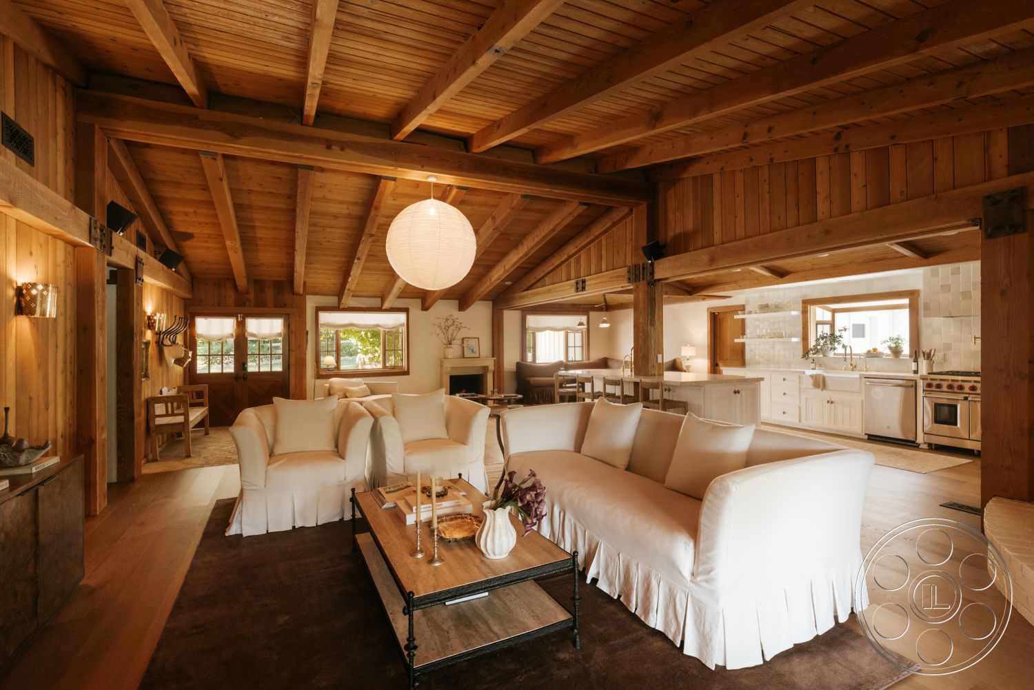 Modern-Farmhouse 20 - wood coffee table, cozy seating area, modern pendant lighting, open concept living space, exposed wood ceiling beams, stone fireplace wall, wood paneling walls, large windows providing natural light, hardwood flooring in living area, rustic interior design, contemporary farmhouse style, white slipcovered sofa, white cabinetry in kitchen, vaulted ceiling with timber trusses