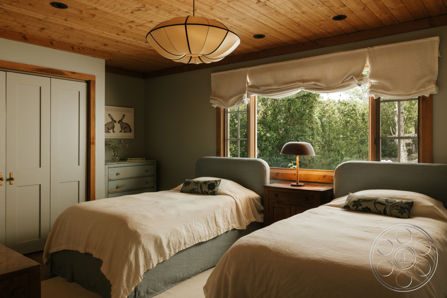 Modern-Farmhouse 20 - wooden bedside table, wood ceiling, exposed wood beams, vaulted ceiling, natural light from windows, wall art with nature theme, glass window treatments, neutral carpet flooring, textured wall paint, wooden window trim, soft beige bedding, dual twin beds, decorative lamp with shade, pastel-colored dresser, green foliage view from window