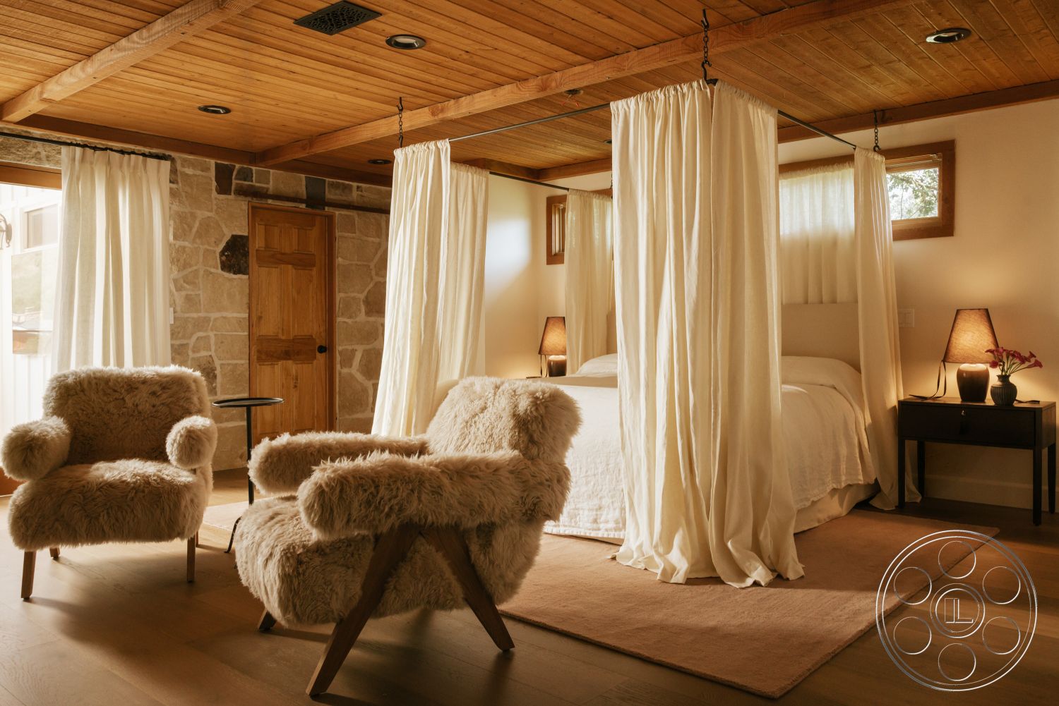 Modern-Farmhouse 20 - stone accent wall, floor-to-ceiling windows, natural wood door, ambient lighting fixtures, exposed wood ceiling beams, neutral painted walls, wooden floorboards, minimalist side table, canopy bed with sheer curtains, fluffy upholstered chairs