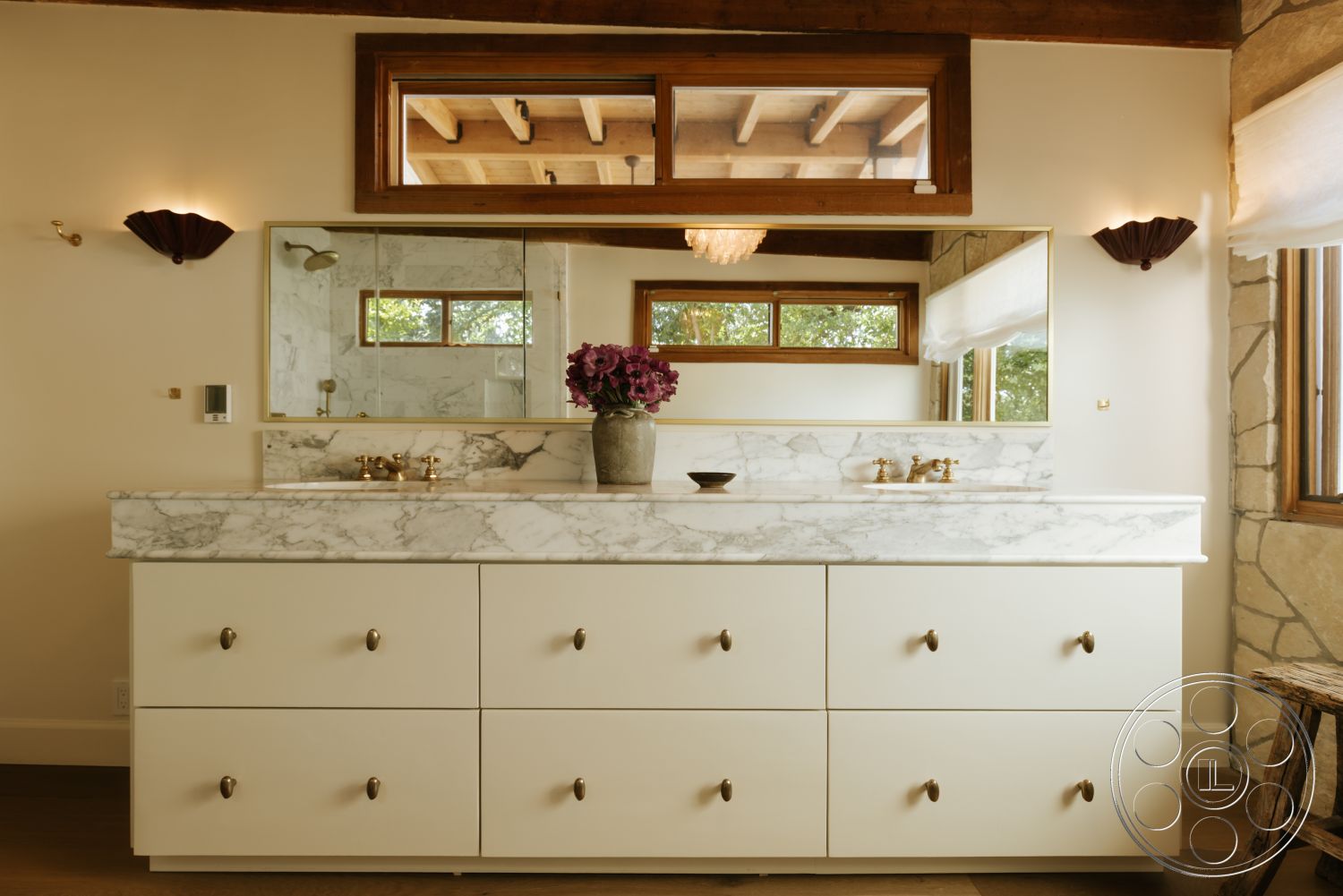 Modern-Farmhouse 20 - stone accent wall, window with natural light, wall-mounted sconces, wooden floor tiles, marble vanity countertop, light-colored wall paint, white cabinetry with brass handles, wooden beams ceiling, floating bathroom sink, rectangle mirror above vanity, plush floral centerpiece