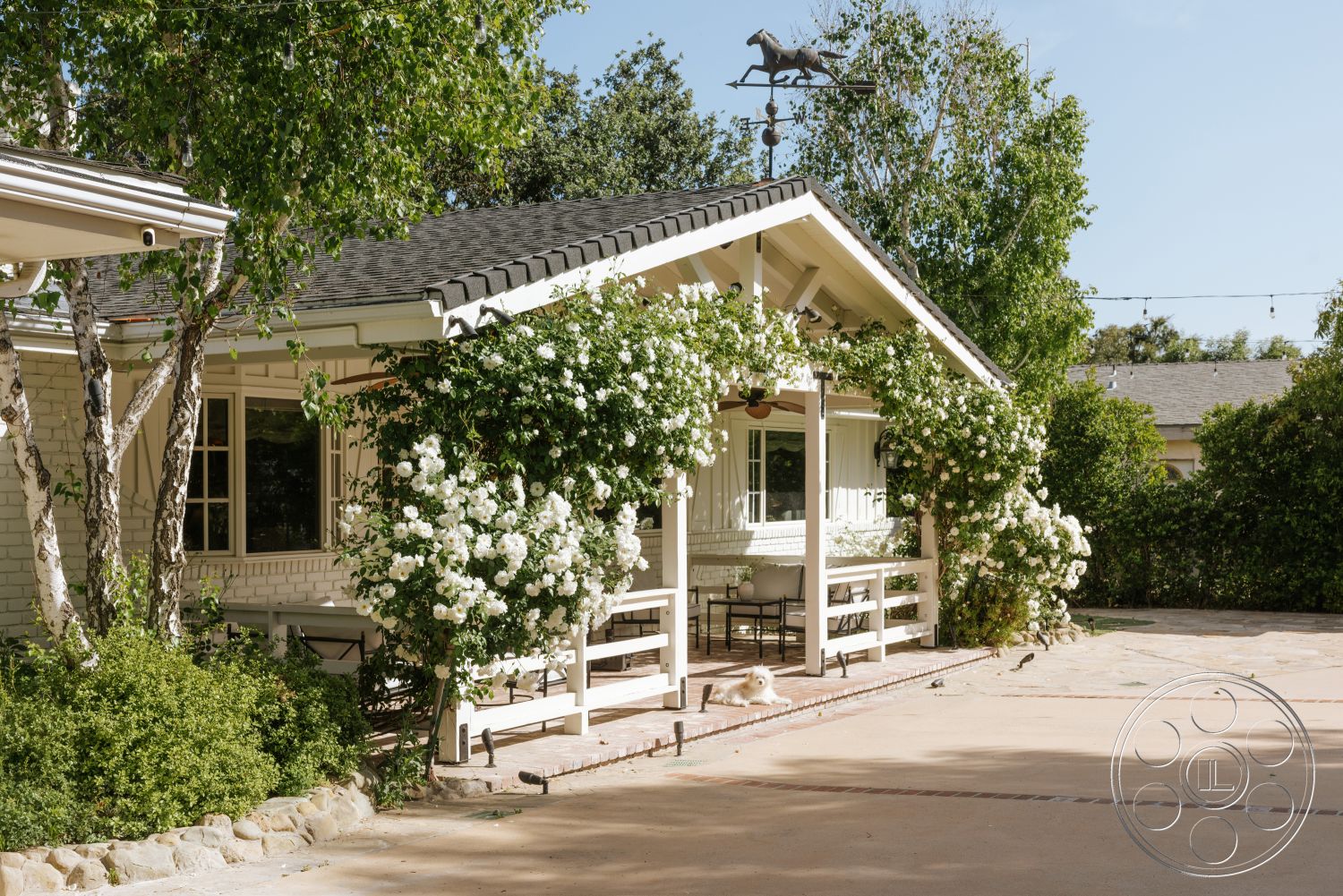 Modern-Farmhouse 20 - exterior, paved driveway, surrounding trees, single-story home, gabled roof, outdoor dining space, white brick walls, flagstone pathway, landscaped garden beds, natural stone garden accents, wooden porch railing, structural wood beams, floral climbing vines, cedar shake roofing, decorative weather vane, covered porch area