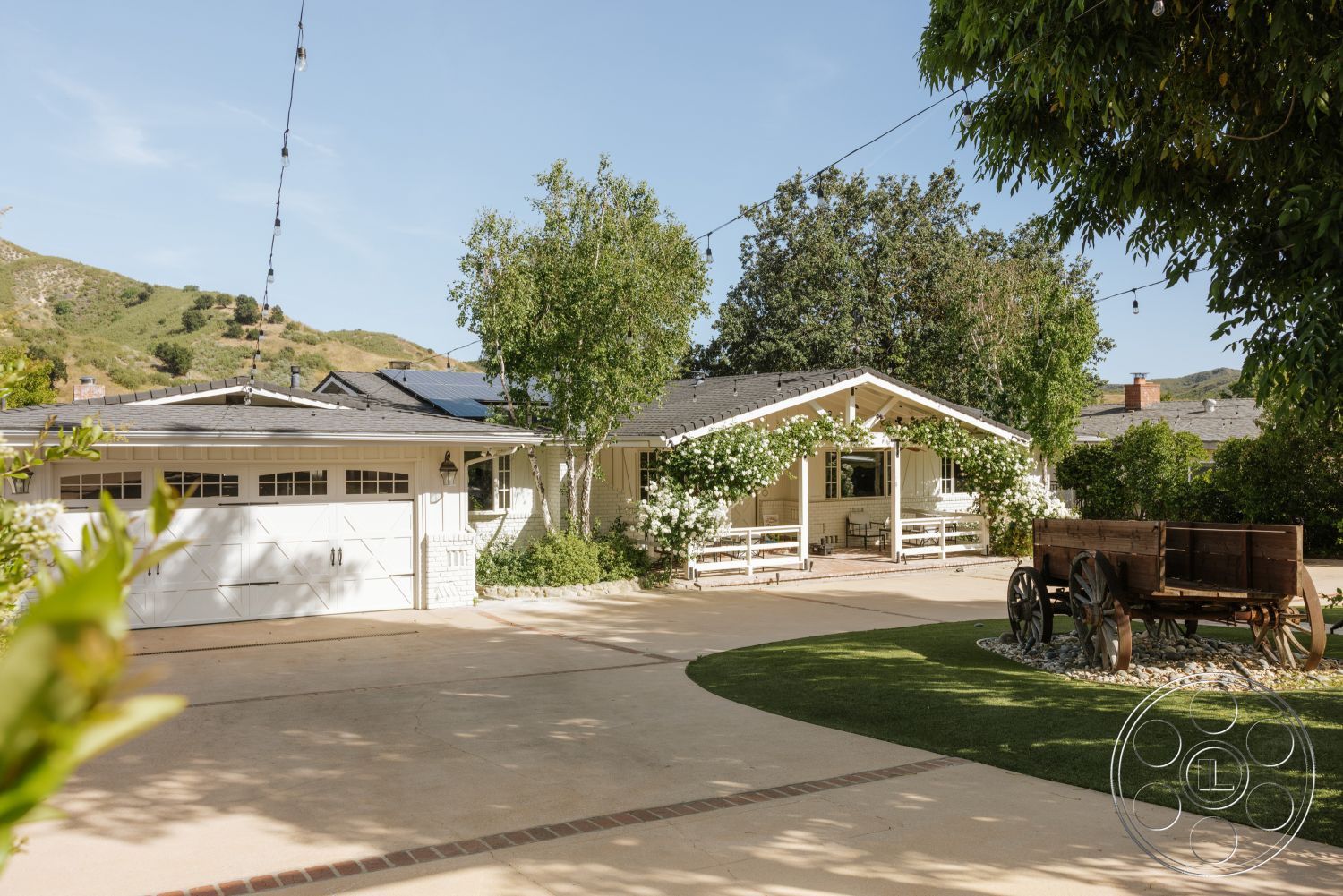 Modern-Farmhouse 20 - exterior, white painted walls, ranch-style home, landscaped yard with grass, mountain landscape view, gray asphalt shingles roof, concrete driveway with brick border, covered front porch with white railing, mixed greenery with flowering plants, wooden vintage cart, string lights above outdoor area