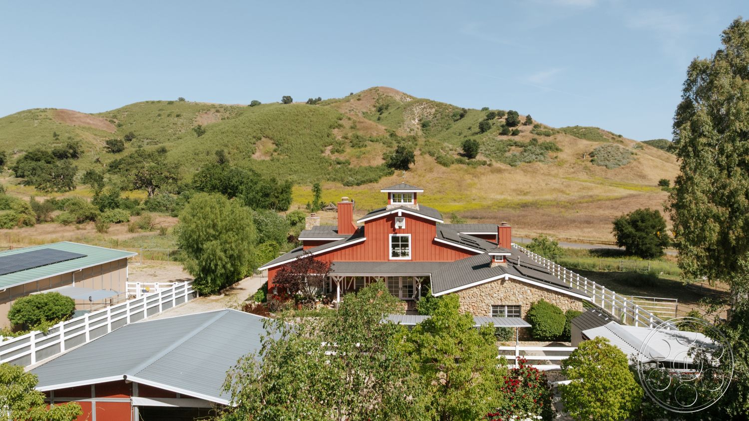 Modern-Farmhouse 20 - large windows, exterior, white wooden fence, metal roof, porch area, outdoor patio space, stone feature wall, red wood siding, gabled roof shape, natural hillside setting, landscaped garden areas, surrounded by rolling hills, country farmhouse style, multiple rooflines
