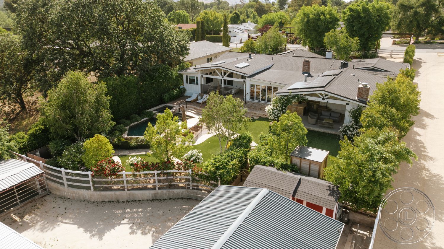 Modern-Farmhouse 20 - exterior, outdoor seating area, surrounding trees, white wooden fence, white wood siding, large floor-to-ceiling windows, gravel driveway, green grass lawn, rectangular pool, garden paths, single-story ranch-style home, landscaped garden beds, concrete pool border, gray asphalt shingle roof, wood deck patio area