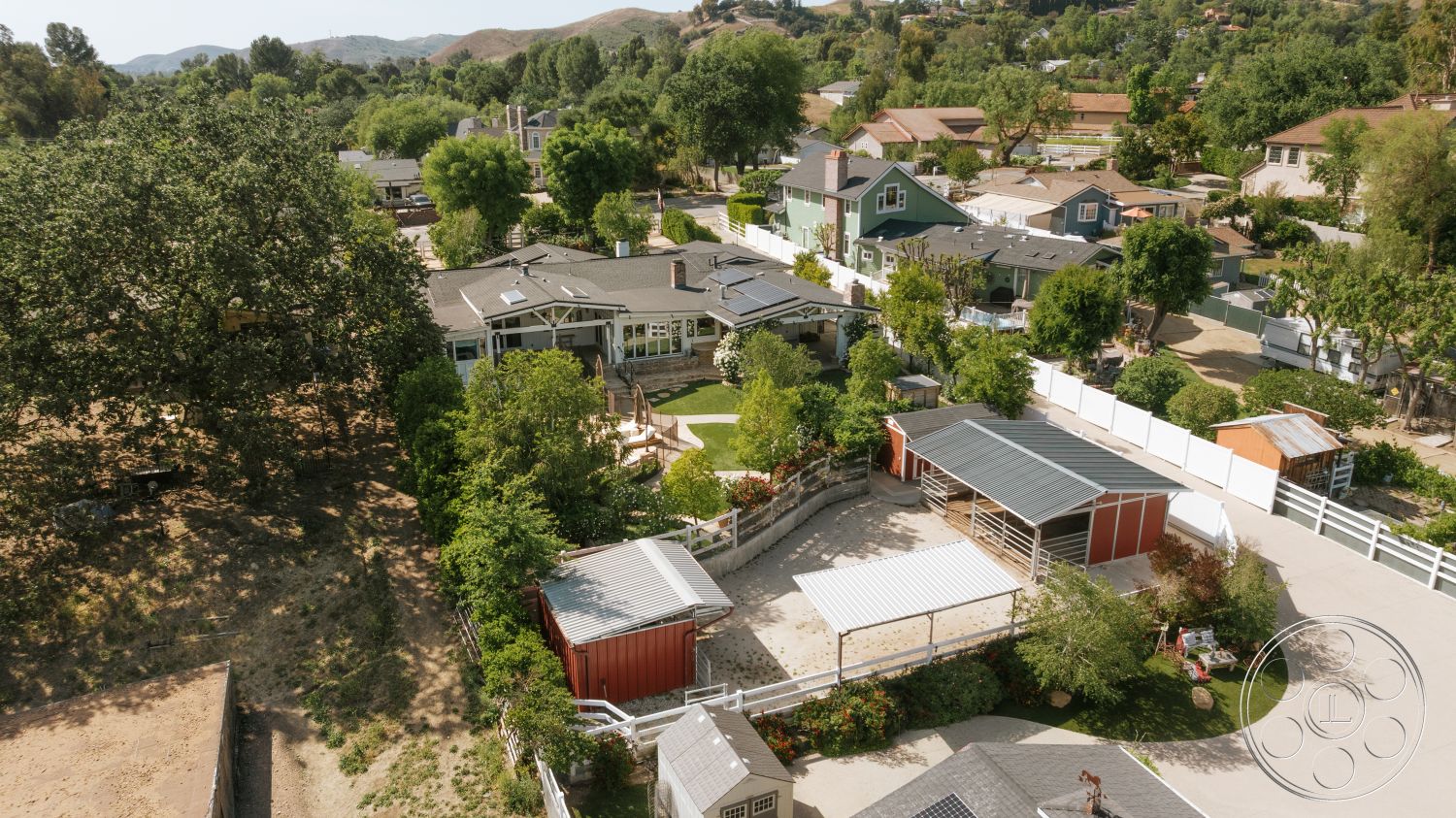 Modern-Farmhouse 20 - exterior, paved driveway, outdoor play area, gray asphalt roof, single-story ranch-style home, mature oak tree, wood deck patio, surrounding hillside location, landscaped garden with shrubs, residential neighborhood setting, metal roof structure, white vinyl fence, red storage barn