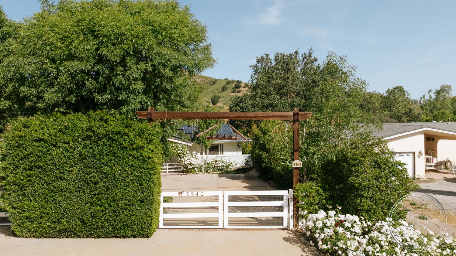 Modern-Farmhouse 20 - single-story home, concrete driveway, white picket fence, gravel area, hedge landscaping, wooden gate entrance, sloped hillside setting, flower bed with white roses, vertical wooden posts, greenery and trees surrounding property, natural light exterior