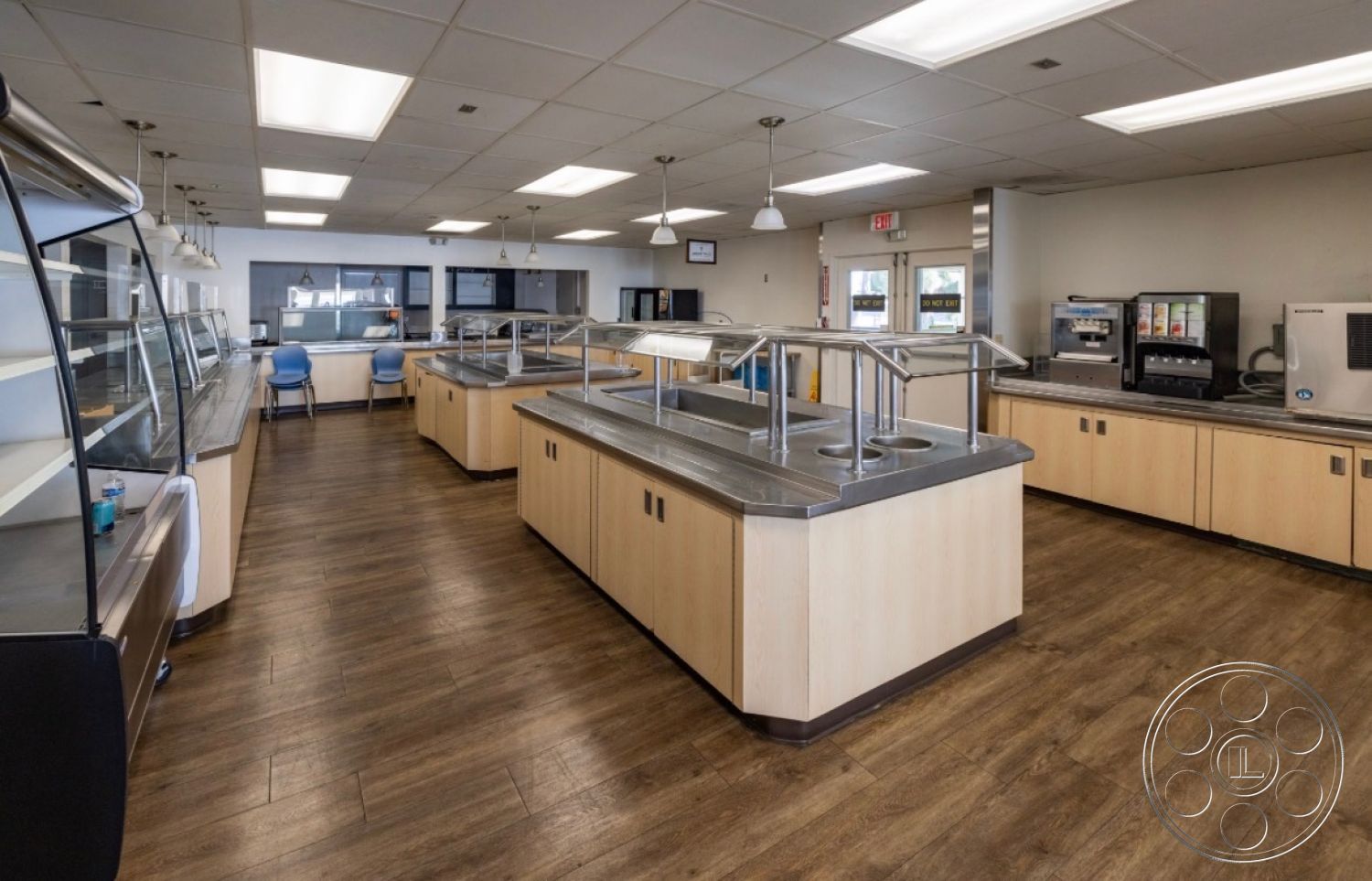 School 1 - large windows, open layout, wood cabinetry, drop ceiling, laminate wood flooring, glass display cases, flush mount ceiling lights, industrial pendant lighting, stainless steel counters, metal serving stations, blue seating, contemporary cafeteria style
