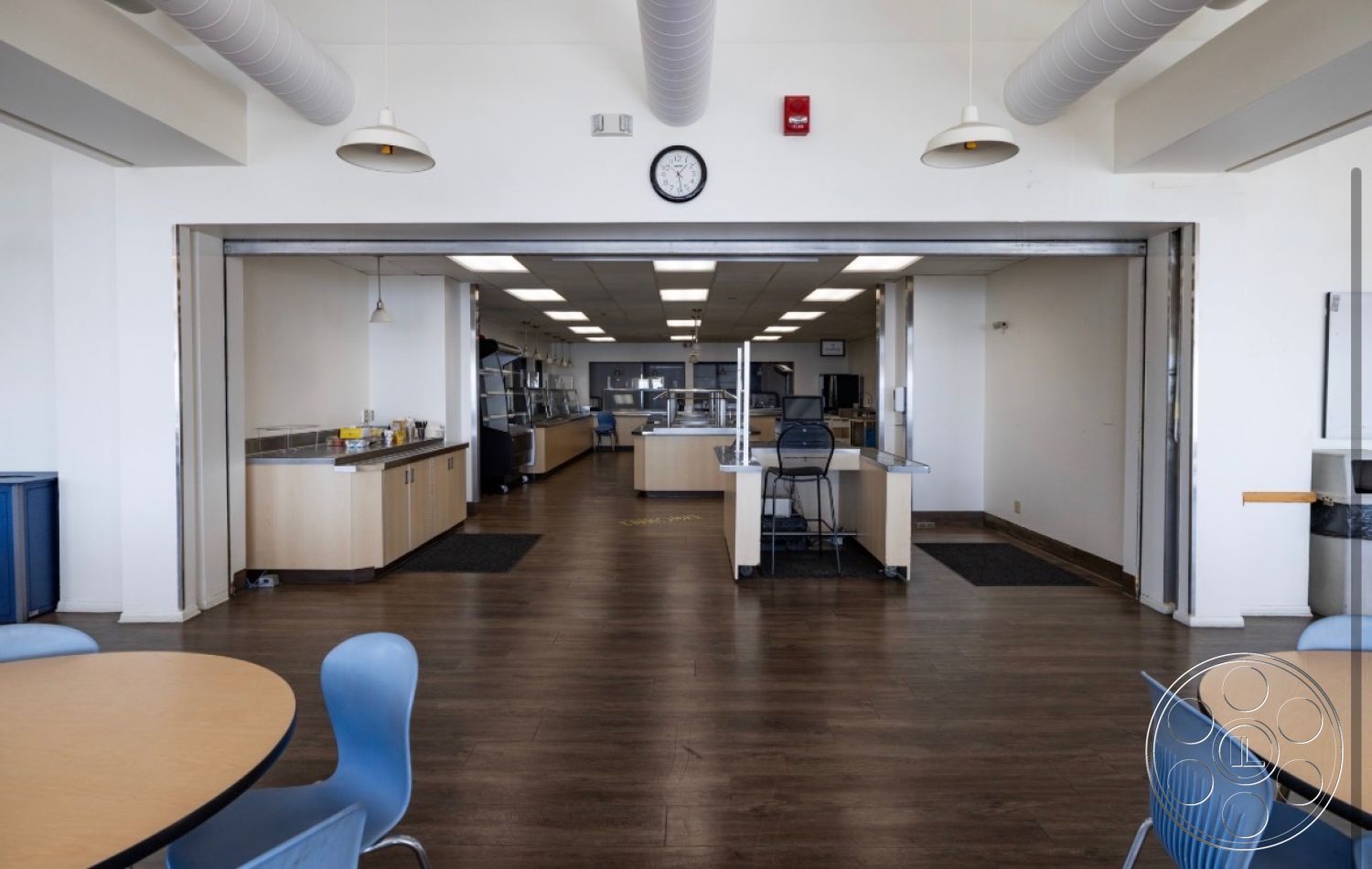 School 1 - modern pendant lighting, floor-to-ceiling windows, interior dining area, open kitchen layout, white painted walls, wood laminate flooring, round wooden dining tables, commercial kitchen area, stainless steel serving counters, blue plastic dining chairs, drop ceiling with ventilation ducts, large pass-through opening, yellow floor markings