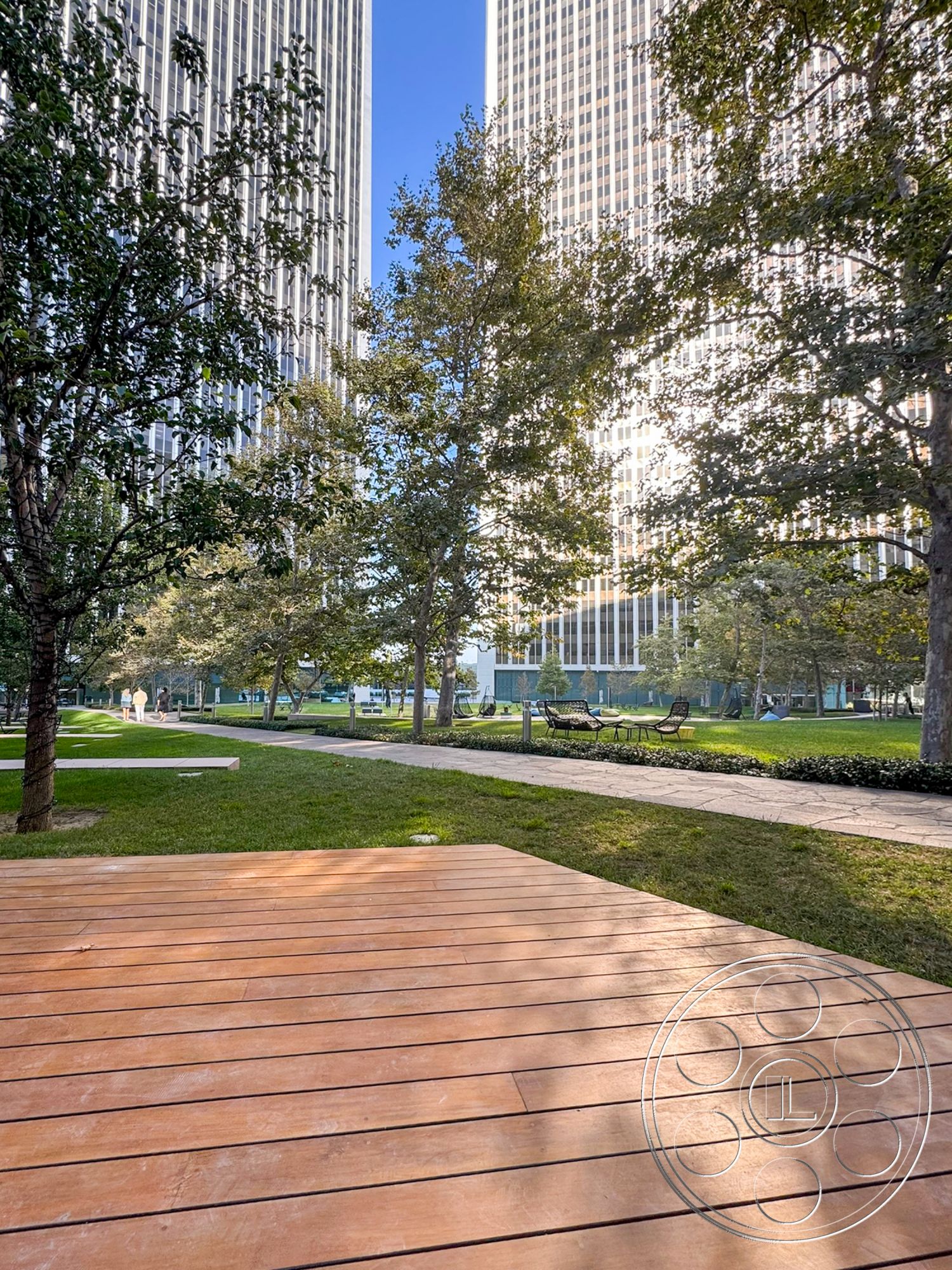 Ave Of The Stars - urban environment, planted trees, wood deck patio, landscaped green lawn, modern skyscraper exterior, gravel walking path, public park setting, sunny day atmosphere