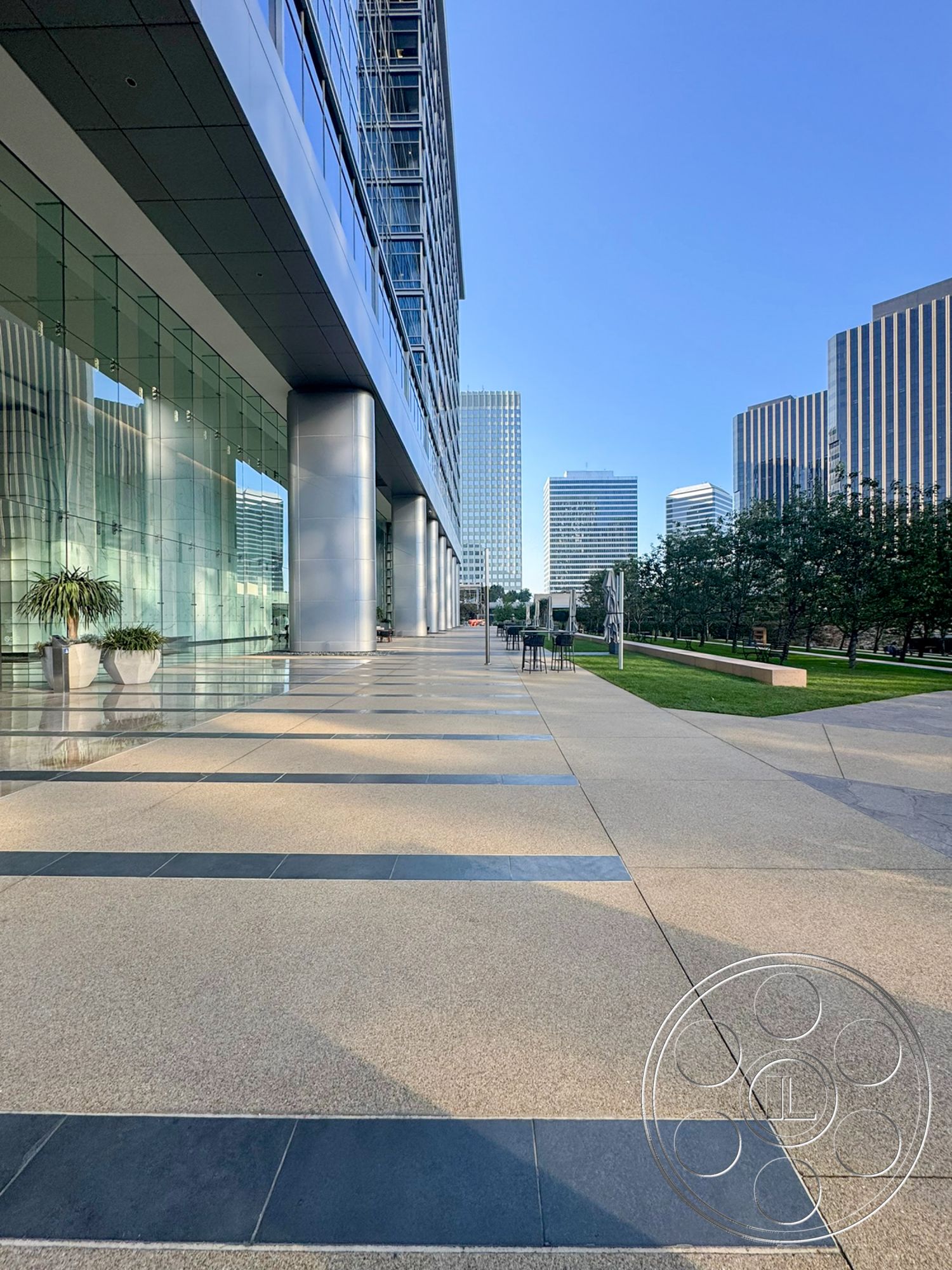 Ave Of The Stars - exterior, outdoor seating area, landscaped green area, city skyline view, modern urban setting, steel structural columns, glass curtain wall facade, concrete plaza flooring, textured concrete walkway, rectangular potted plants, horizontal band of blue glass