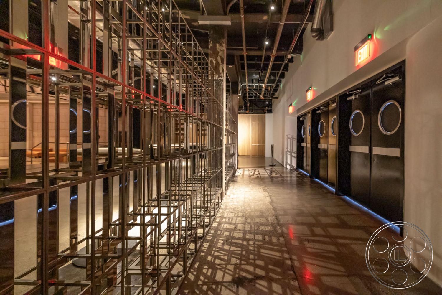 Sunset Campus - polished concrete flooring, minimalist design aesthetics, open layout space, industrial-style hallway, black metal doors with circular windows, exposed metal framework interior, direct ceiling lighting with exposed beams, flush-mounted exit signs, colorful LED accent lighting