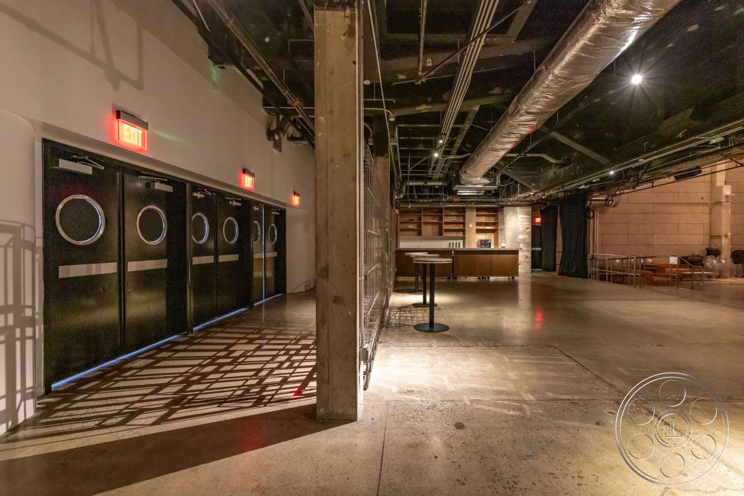 Sunset Campus - polished concrete floor, minimalist design elements, concrete walls, urban setting, dark color palette, wooden bar counter, ambient lighting fixtures, industrial-style interior, exposed concrete flooring, black metal exit doors, open ceiling with exposed ductwork, floor-to-ceiling shelving, architectural shadows on floor, commercial venue interior