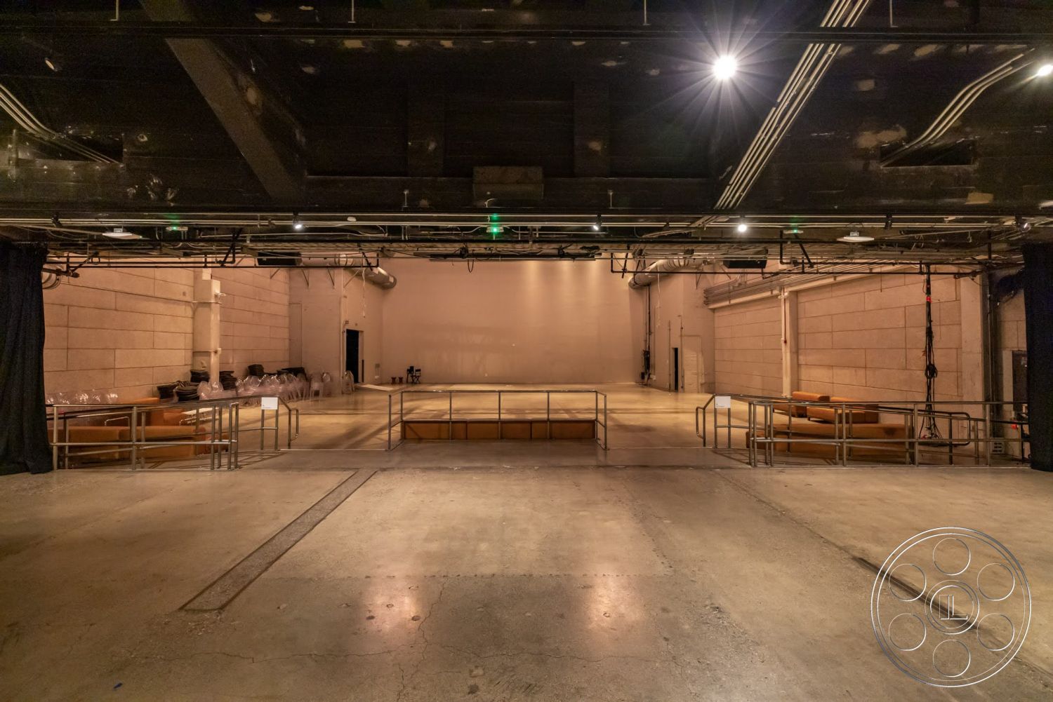 Sunset Campus - textured concrete walls, exposed concrete floor, circular lighting fixtures, industrial architectural style, black ceiling with exposed beams, open performance space, theater stage area, interior venue setting, seating area with modern brown sofas