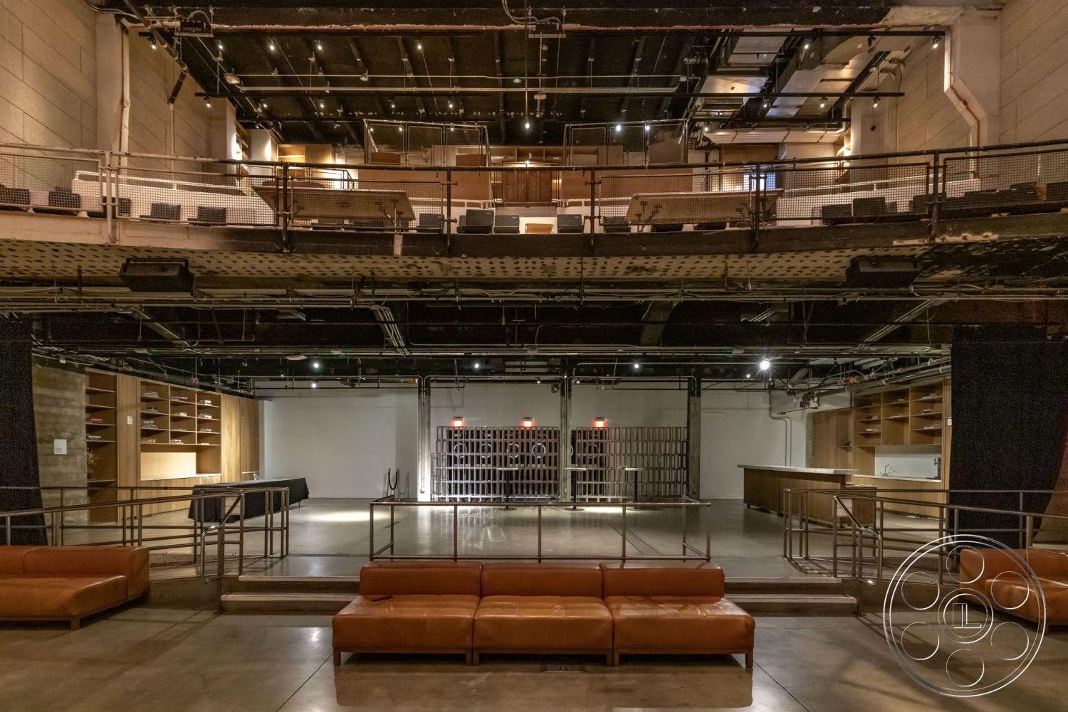 Sunset Campus - open floor plan, concrete flooring, exposed concrete walls, industrial lighting fixtures, metal railing details, black acoustic ceiling panels, wood shelving units, theater-style seating layout, leather lounge seating, bar area with wood cabinetry