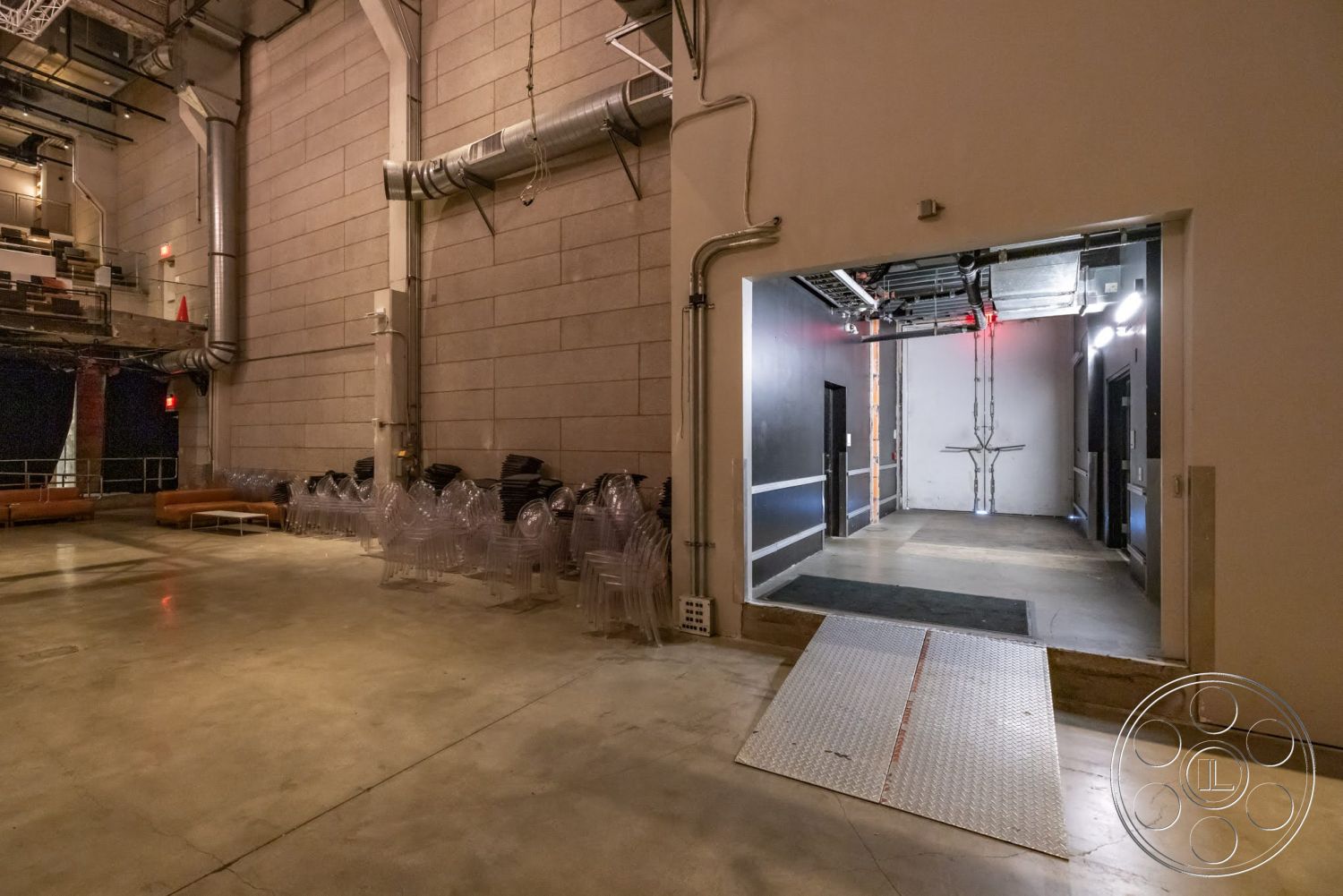 Sunset Campus - concrete floor, exposed concrete walls, glass entry door, black painted walls, acoustic ceiling tiles, steel support beams, industrial ceiling, metal loading dock ramp, seating area with modular couches, clear plastic stacking chairs, theater space