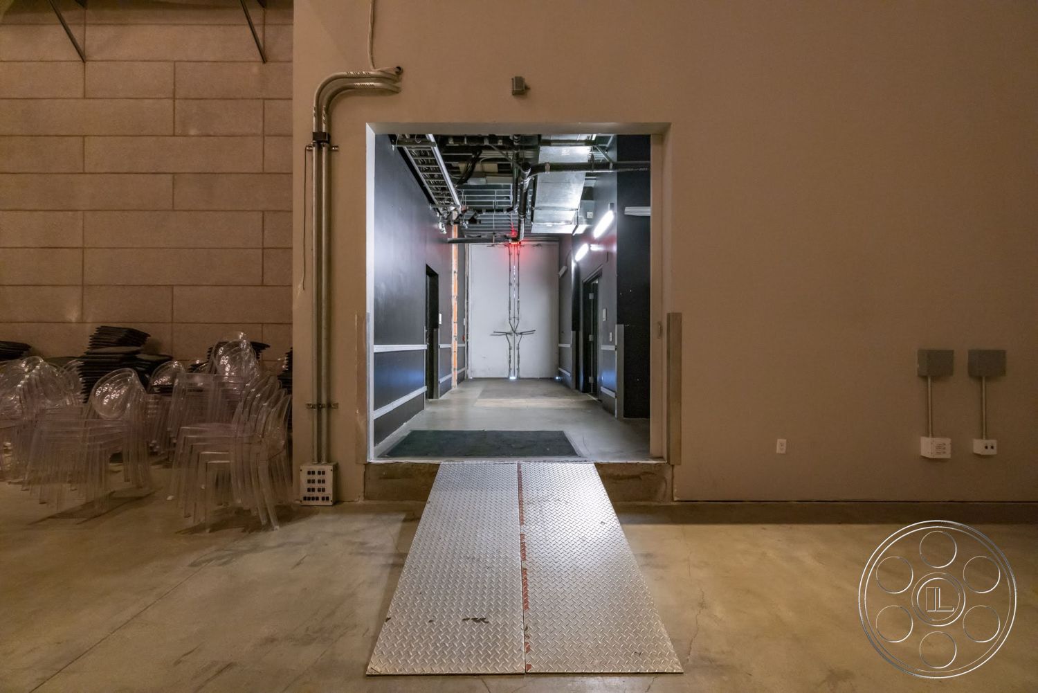 Sunset Campus - minimalist design, interior space, dim ambient lighting, exposed concrete floor, large entryway, gray painted walls, metal loading ramp, industrial-style walls, high ceiling with exposed piping, stacked acrylic chairs