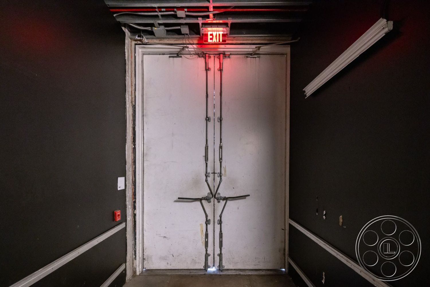Sunset Campus - exposed ceiling structure, black painted walls, interior hallway, fluorescent ceiling lights, wall-mounted fire alarm, cable management, metal exit door, chain lock hardware, emergency exit light, safety exit signage