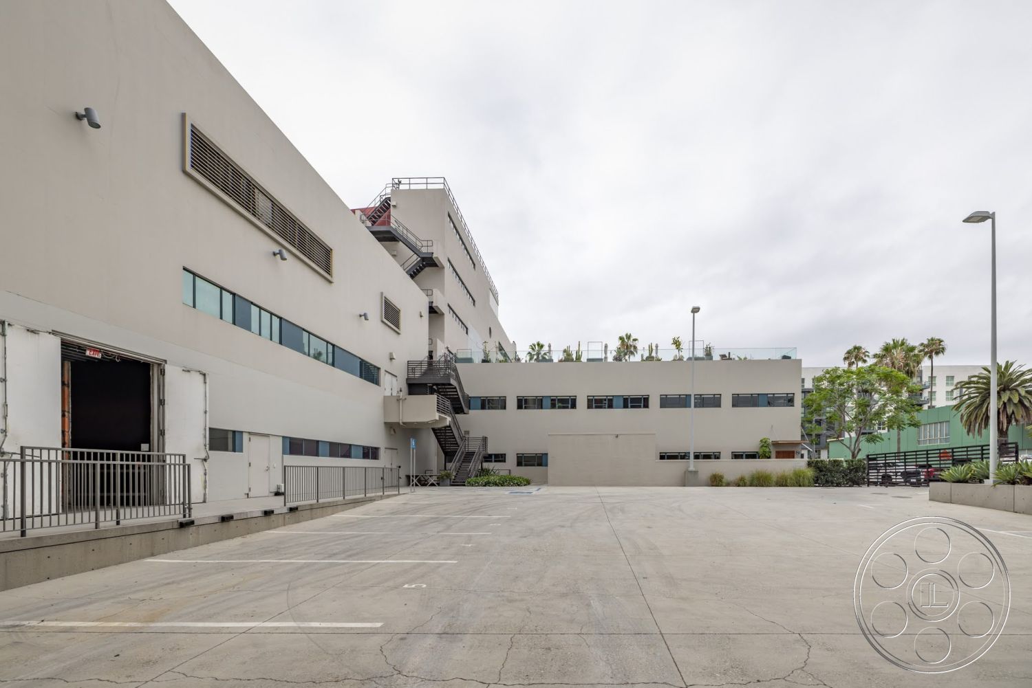 Sunset Campus - exterior, minimalist design elements, urban setting, floor-to-ceiling windows, concrete parking lot, landscaped green space, flat roof design, modern industrial architecture, stucco building façade, steel fire escape staircase, corrugated metal rooftop