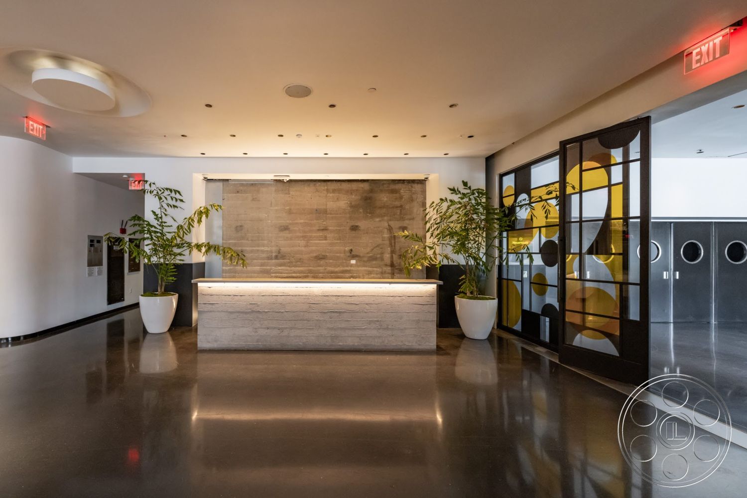 Sunset Campus - neutral color palette, minimalist interior design, floor-to-ceiling glass doors, exposed concrete reception desk, black polished concrete flooring, decorative black metal partition, modern ceiling lighting, textured gray wall treatment, large potted indoor plants, lighted reception counter, open lobby area