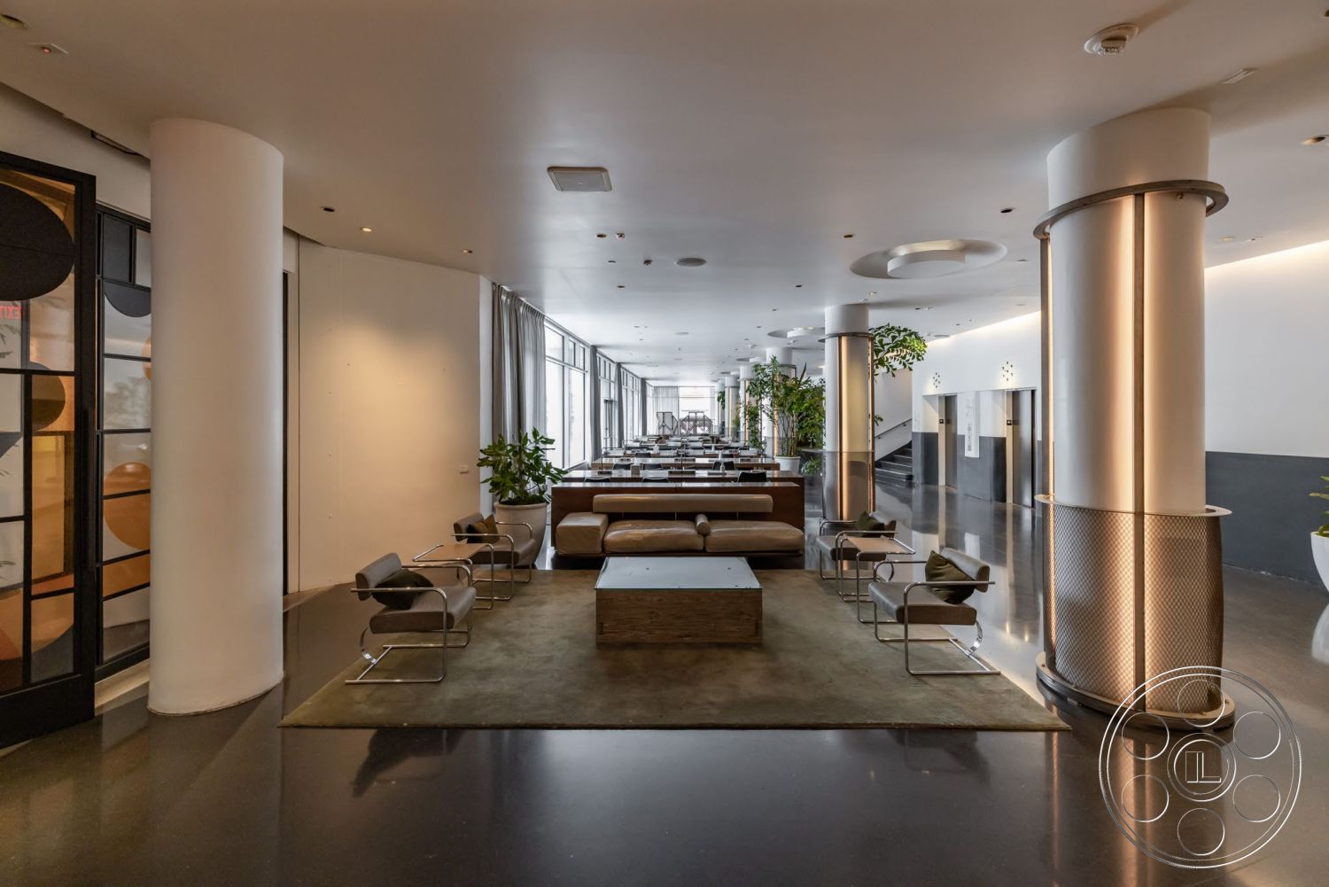 Sunset Campus - neutral color palette, wooden coffee table, polished concrete flooring, green indoor plants, lounge area, modern architectural style, metal stair railings, exposed concrete columns, linear ceiling lighting, fabric lounge chairs, tall windows with drapes, low-profile sofas, area rug in seating area
