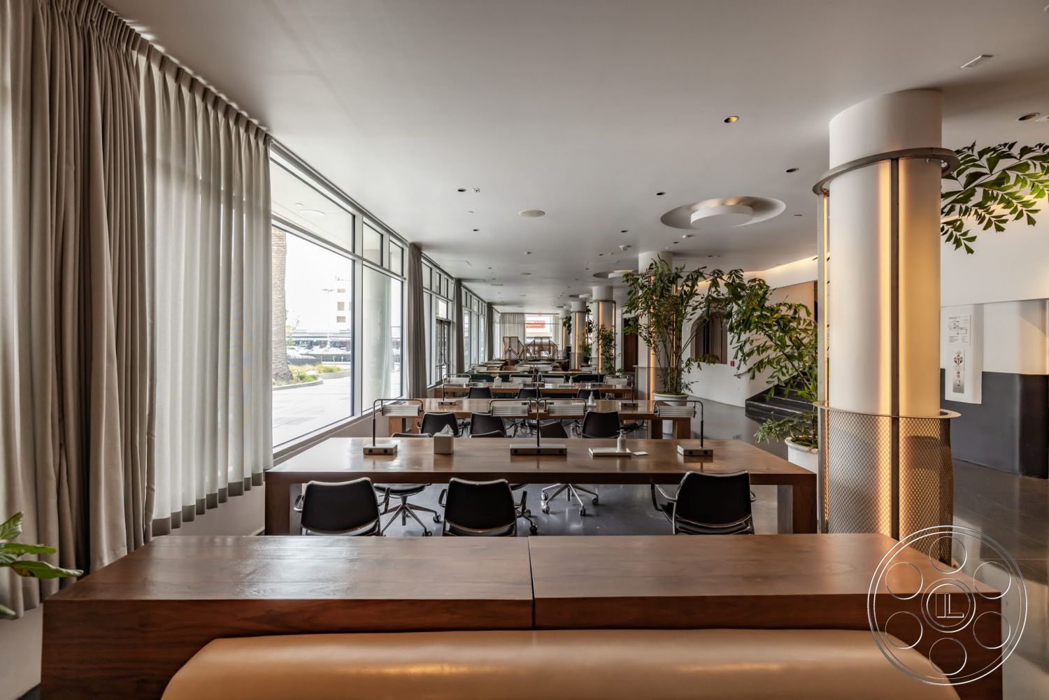 Sunset Campus - concrete floors, floor-to-ceiling windows, industrial lighting fixtures, green indoor plants, neutral color scheme, black office chairs, modern minimalist design, exposed concrete columns, open workspace layout, wood dining tables, beige drapery curtains