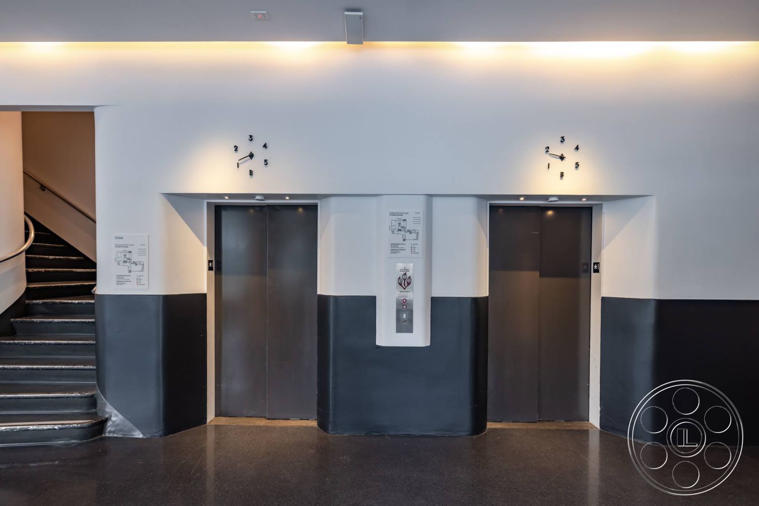Sunset Campus - polished concrete flooring, modern wall clock, circular staircase, white painted walls, stainless steel elevator doors, ambient ceiling lighting, elevator call buttons, black and white wall treatment, glossy black wall base, signage with floor map
