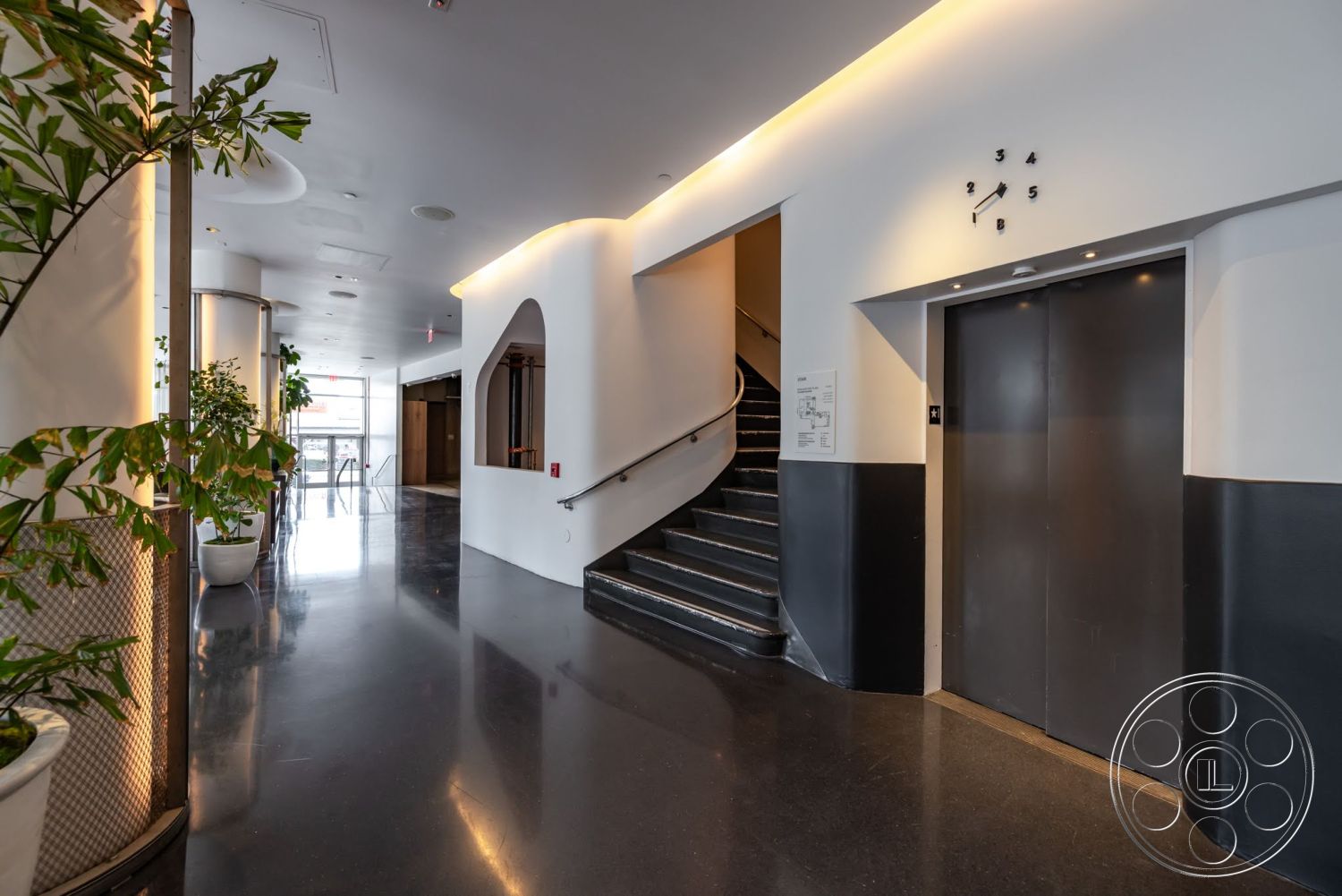 Sunset Campus - polished concrete flooring, black accent wall, recessed ceiling lighting, contemporary design style, open lobby space, modern elevator design, white wall treatments, minimalist architectural elements, curved staircase with black steps, lighted plant display, arced wall opening