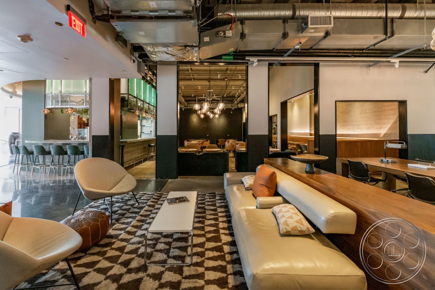 Sunset Campus - polished concrete flooring, geometric area rug, wood accent wall, wooden bar counter, low coffee table, white leather sofa, pendant lighting fixtures, modern lounge seating, exposed ductwork ceiling, open seating area, dark wall paneling, glass bar back display, green upholstered bar stools, artistic accent pillows, lounge area with a view