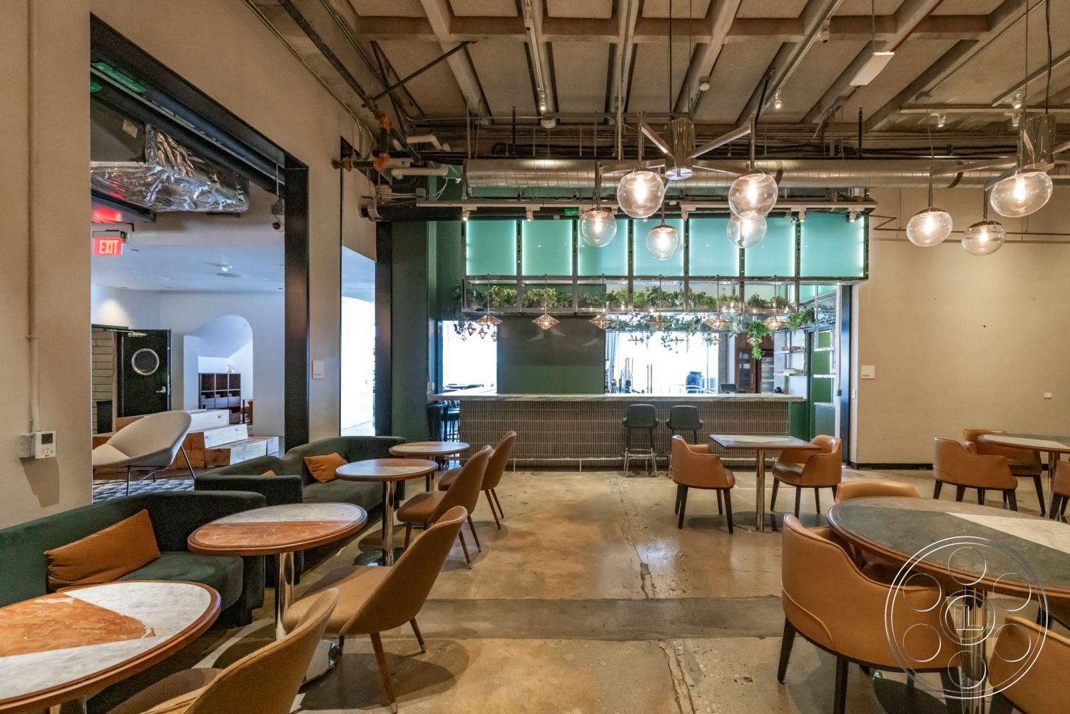 Sunset Campus - minimalist design elements, metal bar counter, open floor plan layout, white painted walls, exposed ductwork ceiling, exposed concrete flooring, industrial styled lighting fixtures, glass bar with green panels, wooden tables with circular tops, upholstered seating with curved backs, urban café interior