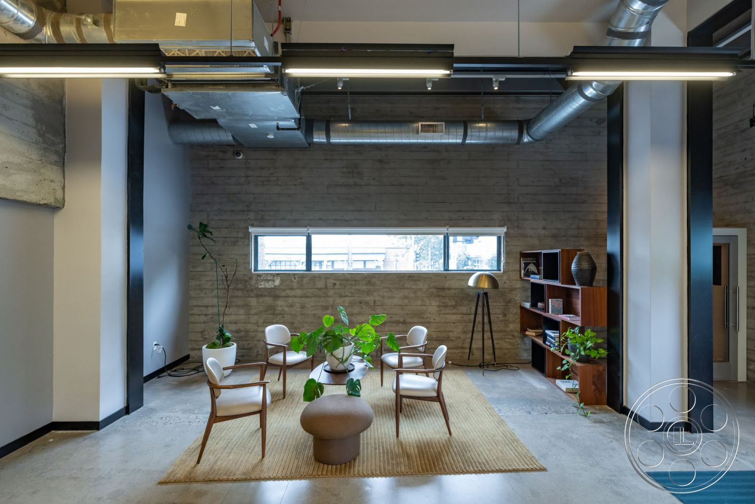 Sunset Campus - polished concrete floor, exposed concrete walls, industrial lighting fixtures, green indoor plants, natural fiber area rug, metal ductwork ceiling, mid-century modern armchairs, wooden frame bookshelves, colorful decorative vases