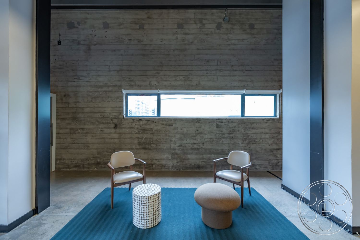 Sunset Campus - minimalist design, textured area rug, concrete flooring, exposed concrete walls, industrial style, mid-century modern furniture, wooden accent chairs, rectangular window, circular ottoman