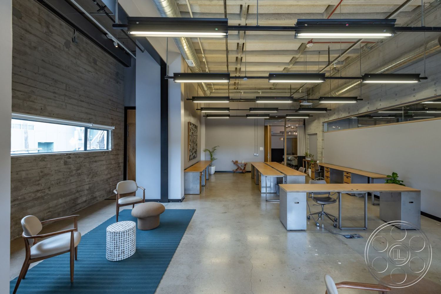 Sunset Campus - neutral color palette, area rug, polished concrete flooring, exposed concrete walls, minimalist furniture, linear pendant lighting, textured wall treatment, comfortable seating area, open office layout, decorative indoor plants, modern office design, office workspace, industrial ceiling fixtures, large horizontal windows, metal desk frames, wood tabletops