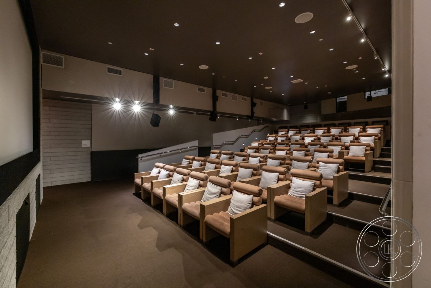 Sunset Campus - recessed lighting, dim ambient lighting, acoustic ceiling panels, projector screen wall, theater room, dark brown carpet flooring, staggered seating arrangement, light wood theater chairs, padded armrests, white throw pillows, concrete block wall accents, built-in audio speakers