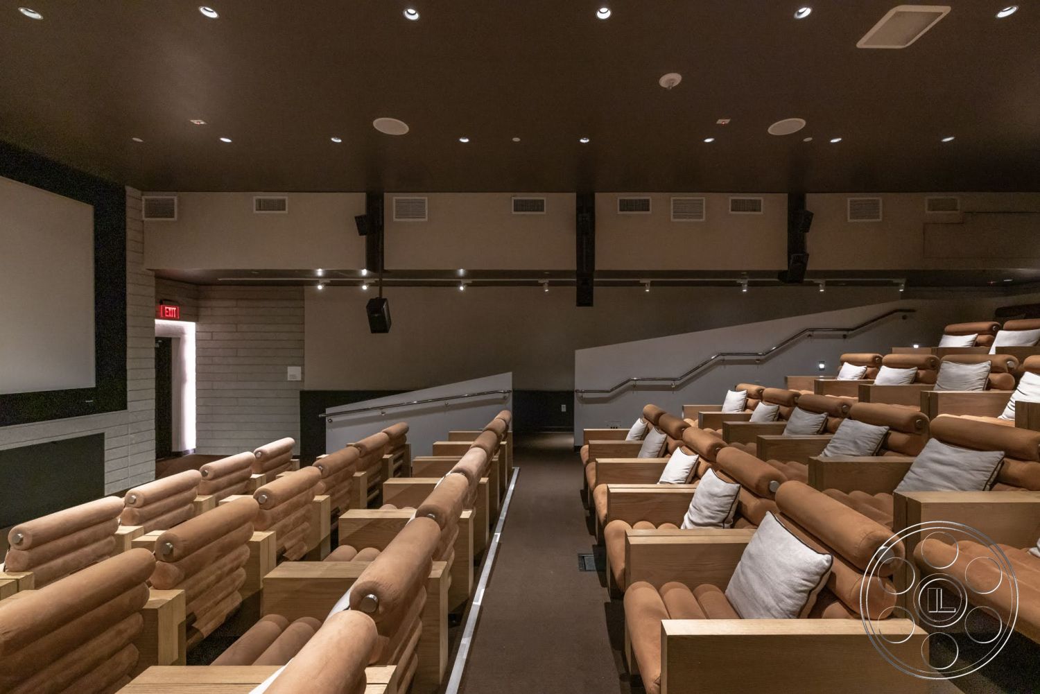 Sunset Campus - dark wood paneling, dim ambient lighting, acoustic wall panels, coffered ceiling design, theater room, reclining theater seats, wood finish theater seating, brown upholstered seating with cushions, soft brown carpet flooring, staircase leading to elevated seating, screening area with projector, side wall mounted speakers
