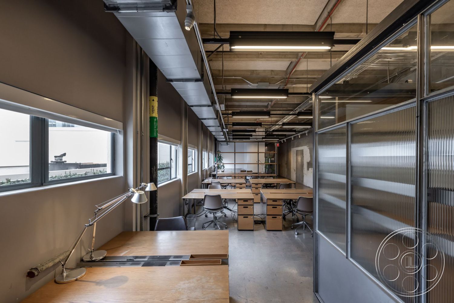 Sunset Campus - concrete floors, industrial lighting fixtures, neutral wall color, exposed ceiling beams, glass partition walls, rectangular workspace layout, potted plants for greenery, large windows with metal frames, wooden desks with storage drawers