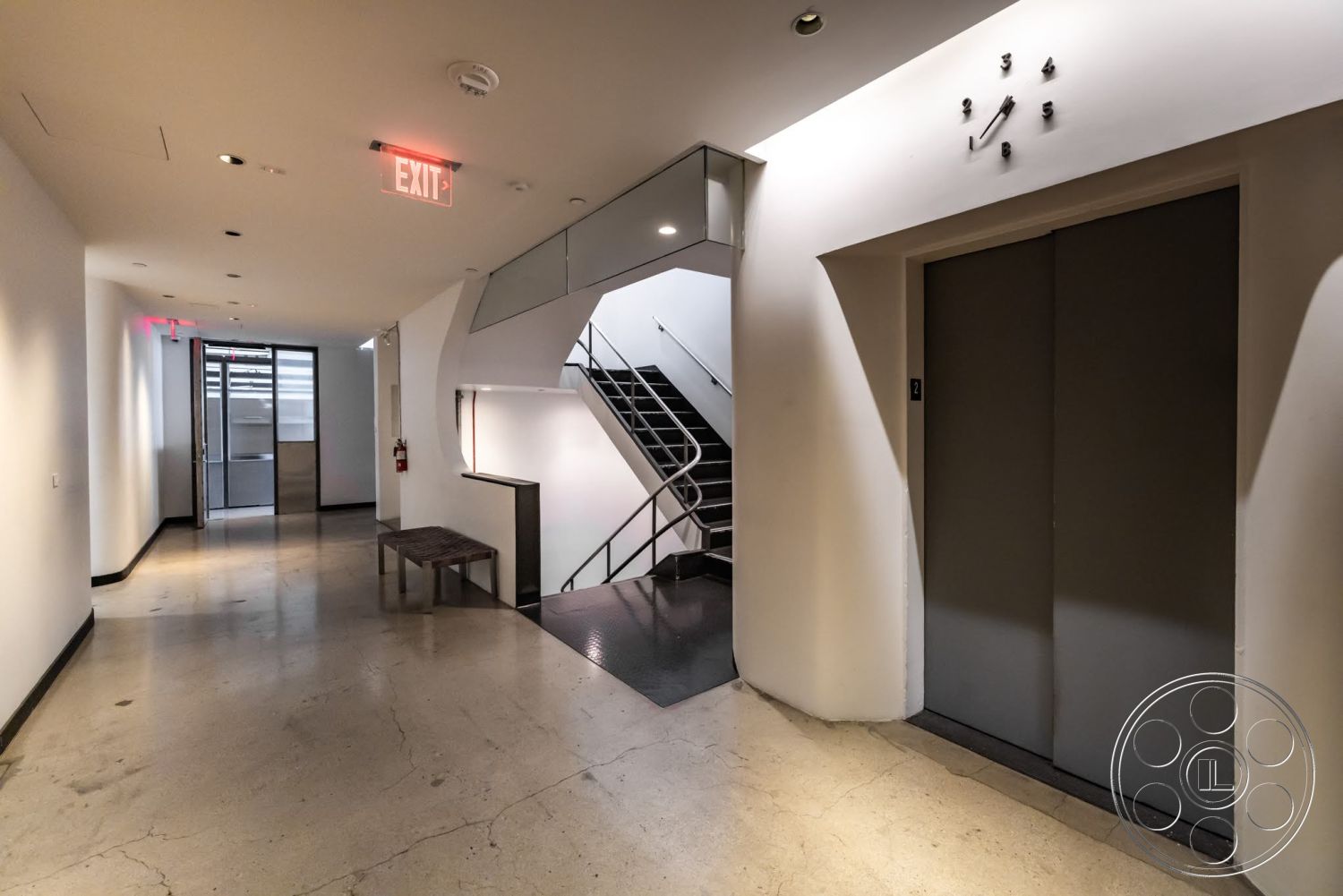 Sunset Campus - polished concrete flooring, exit sign, exposed ceiling lights, interior corridor, glass partition wall, wood bench seating, modern staircase with steel railings, minimalist wall design, single-panel elevator doors, large glass entrance door, curved wall feature