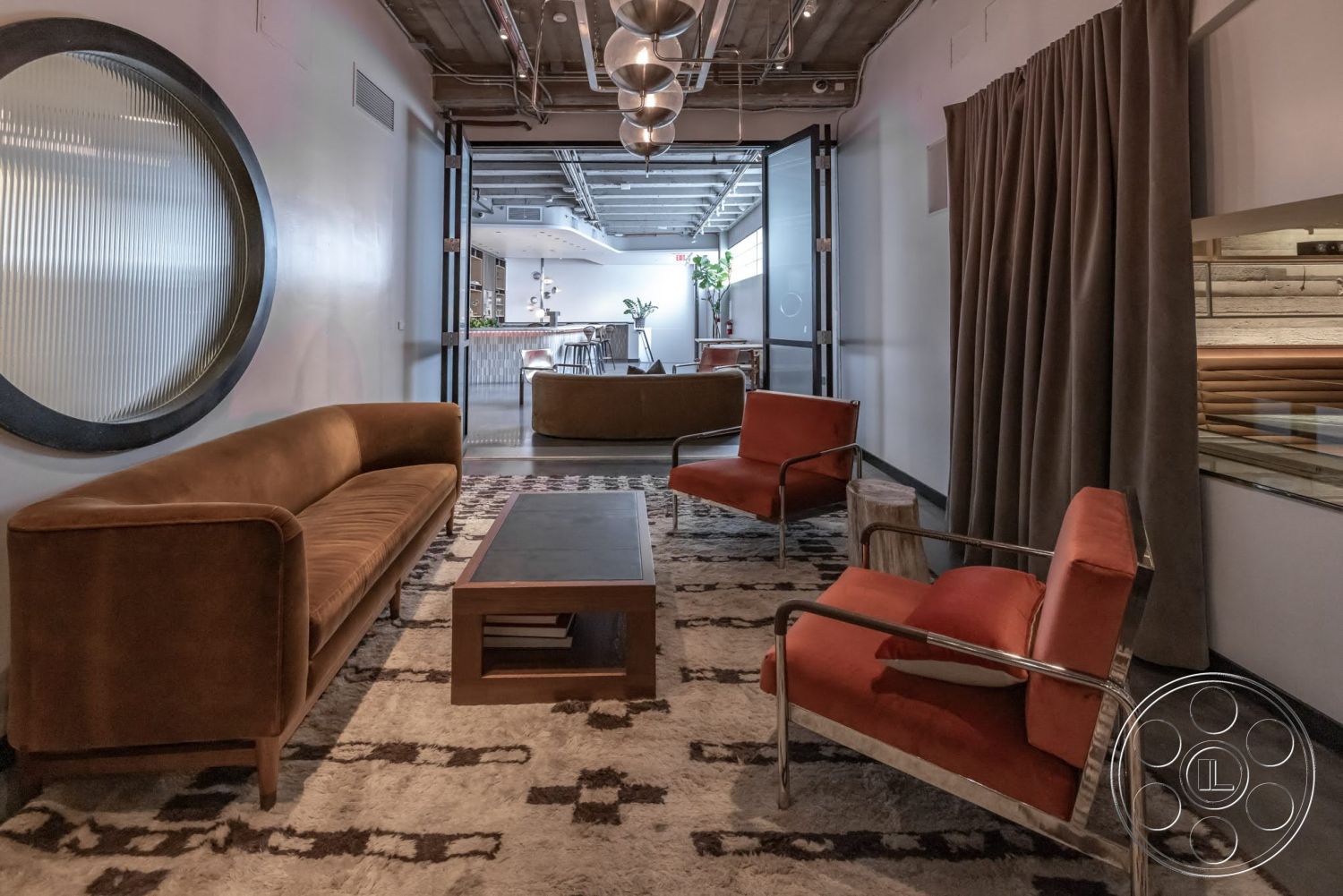 Sunset Campus - neutral color palette, wood coffee table, open floor plan, modern decor elements, geometric area rug, textured wall treatment, exposed concrete ceiling, metal pendant lighting, brown velvet sofa, chrome accent chairs, large round glass window, curtained window treatment