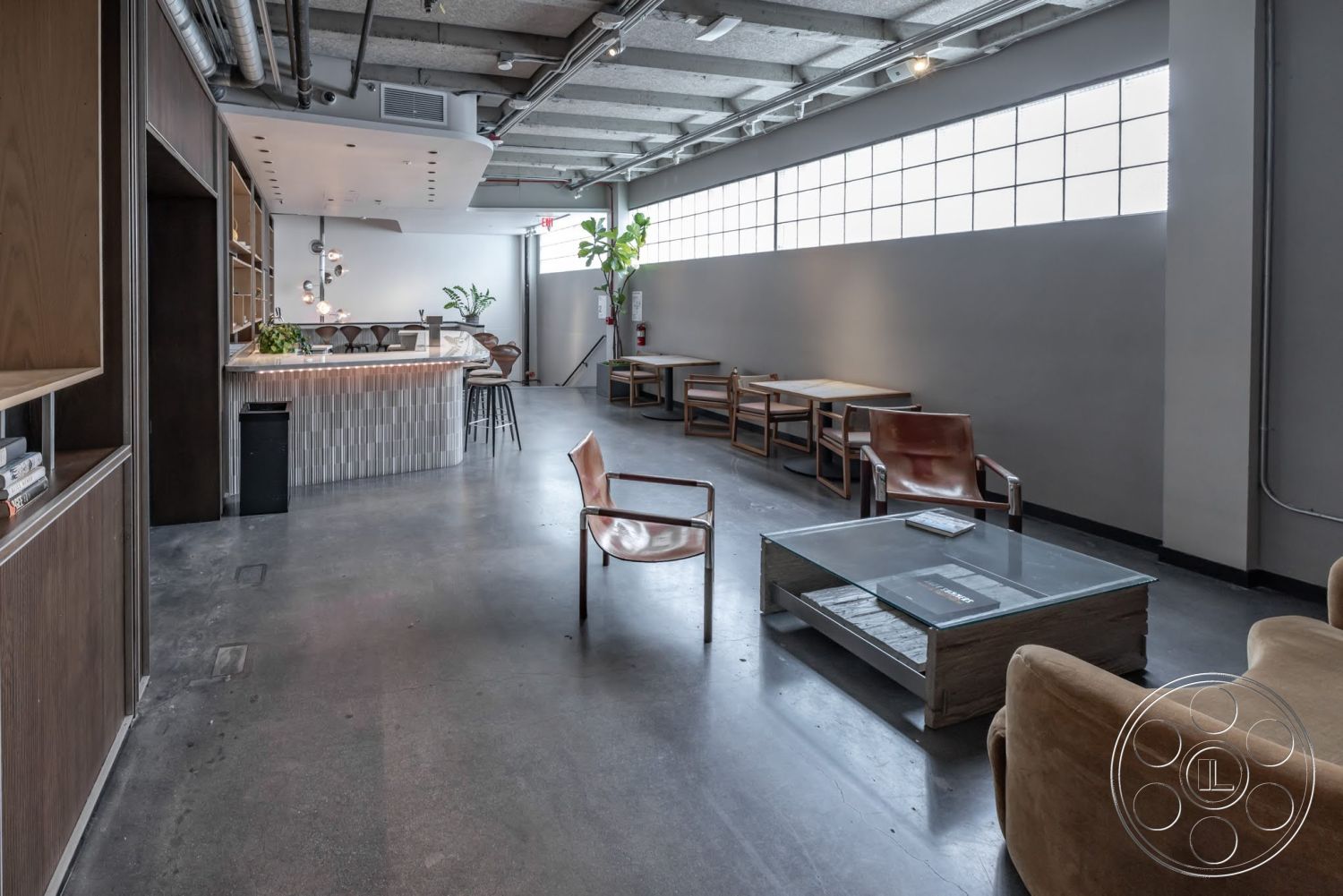 Sunset Campus - polished concrete flooring, exposed concrete walls, urban setting, large industrial windows, glass pendant lighting, rectangular coffee table, minimalist interior design, light wood bar counter, leather lounge chairs, wood shelving units, modern open layout