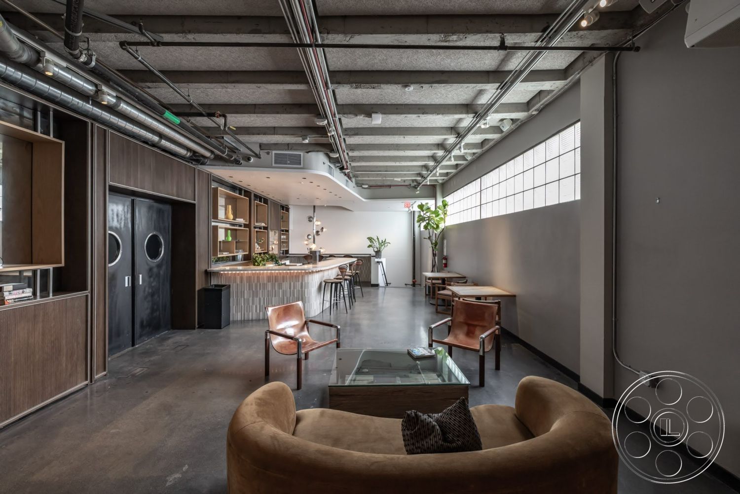 Sunset Campus - minimalist decor, polished concrete floor, glass coffee table, floor-to-ceiling windows, industrial lighting fixtures, green indoor plants, exposed ductwork, bar stools, dark wood paneling, concrete ceiling, open layout space, modern bar with wooden countertop, circular door handles, low-profile seating area, oval lounge chair
